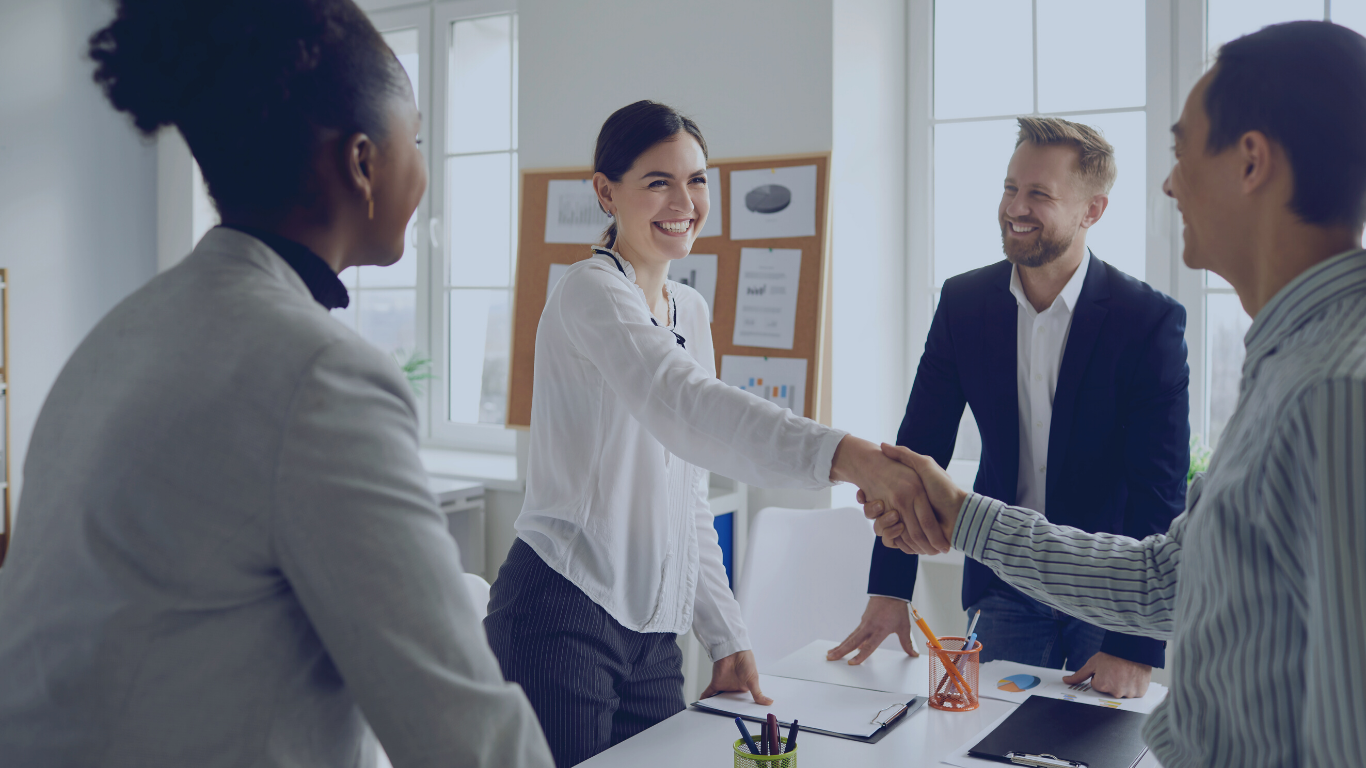 A group of four diverse business professionals in an office, with a woman in a white shirt shaking hands with a man in a striped shirt, while two others, a woman in a gray blazer and a man in a dark suit, look on and smile.