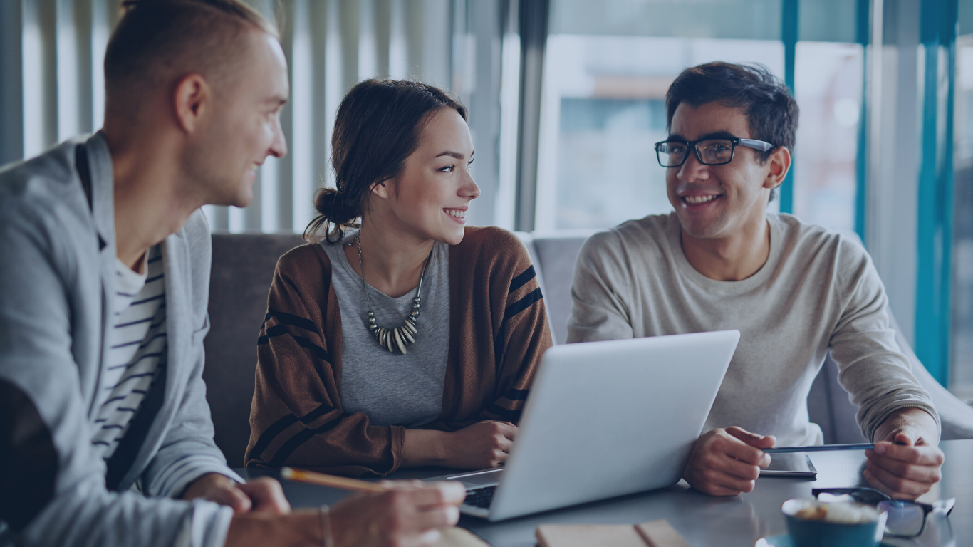 Four young adults sitting at a table with a laptop in a modern, well-lit cafe or office space, smiling and engaging in conversation.