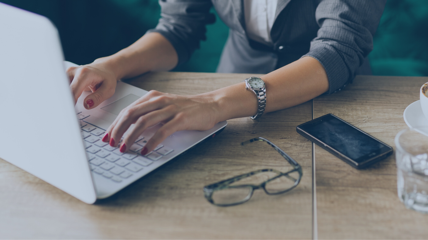 Close-up of a person typing on a laptop at a wooden table, with glasses, a smartphone, and a cup of coffee nearby.