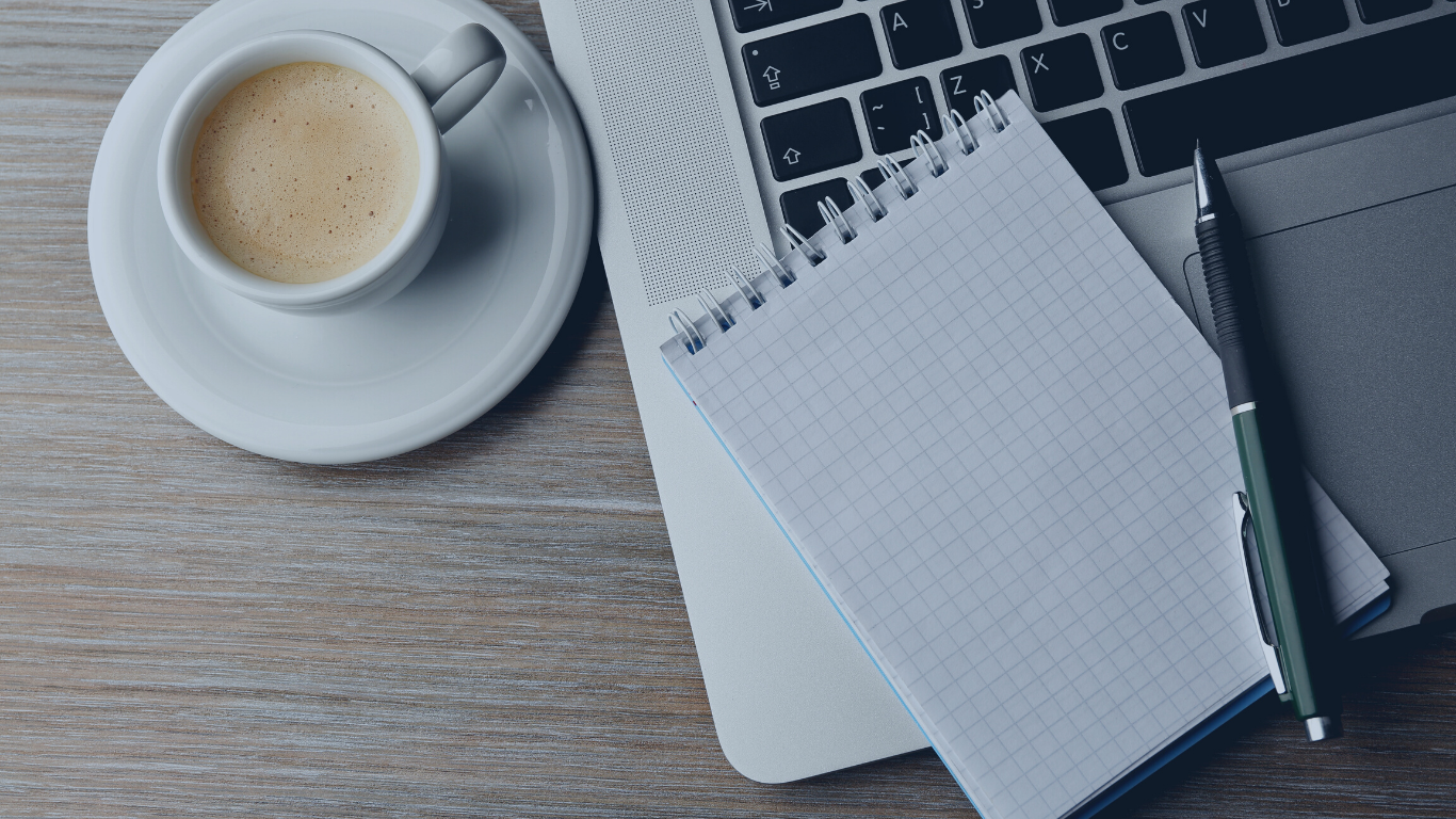 A workspace with a cup of coffee on a saucer, a laptop, a small spiral notepad with graph paper, and a pen on a wooden surface.