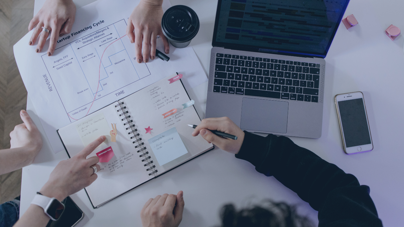 A group of people working at a white table with laptops, smartphones, a coffee cup, and notebooks, discussing finance charts and planning.