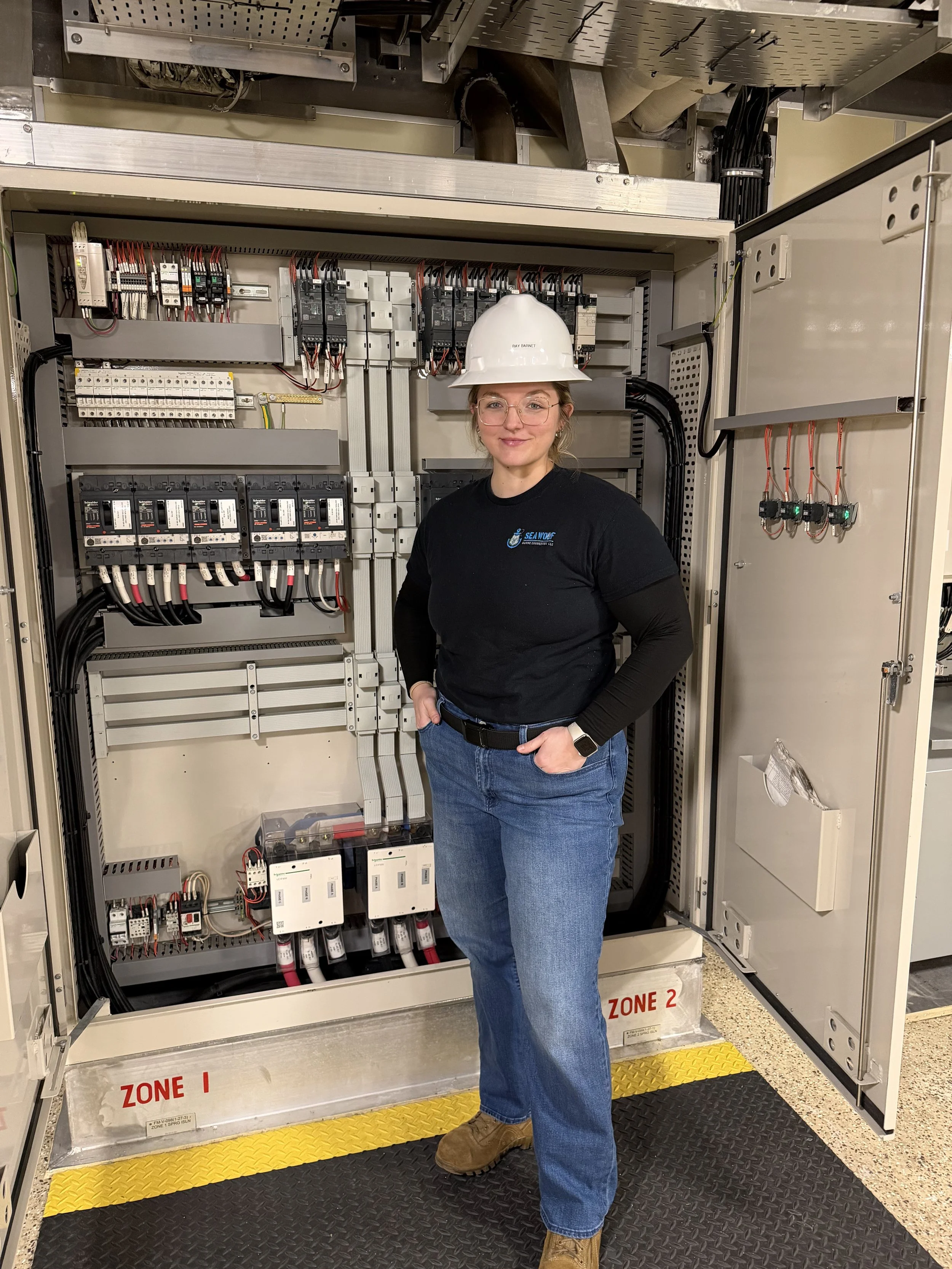 A woman in safety gear, including a white hard hat and glasses, standing in front of an electrical control panel with labeled zones, in an industrial or technical setting.