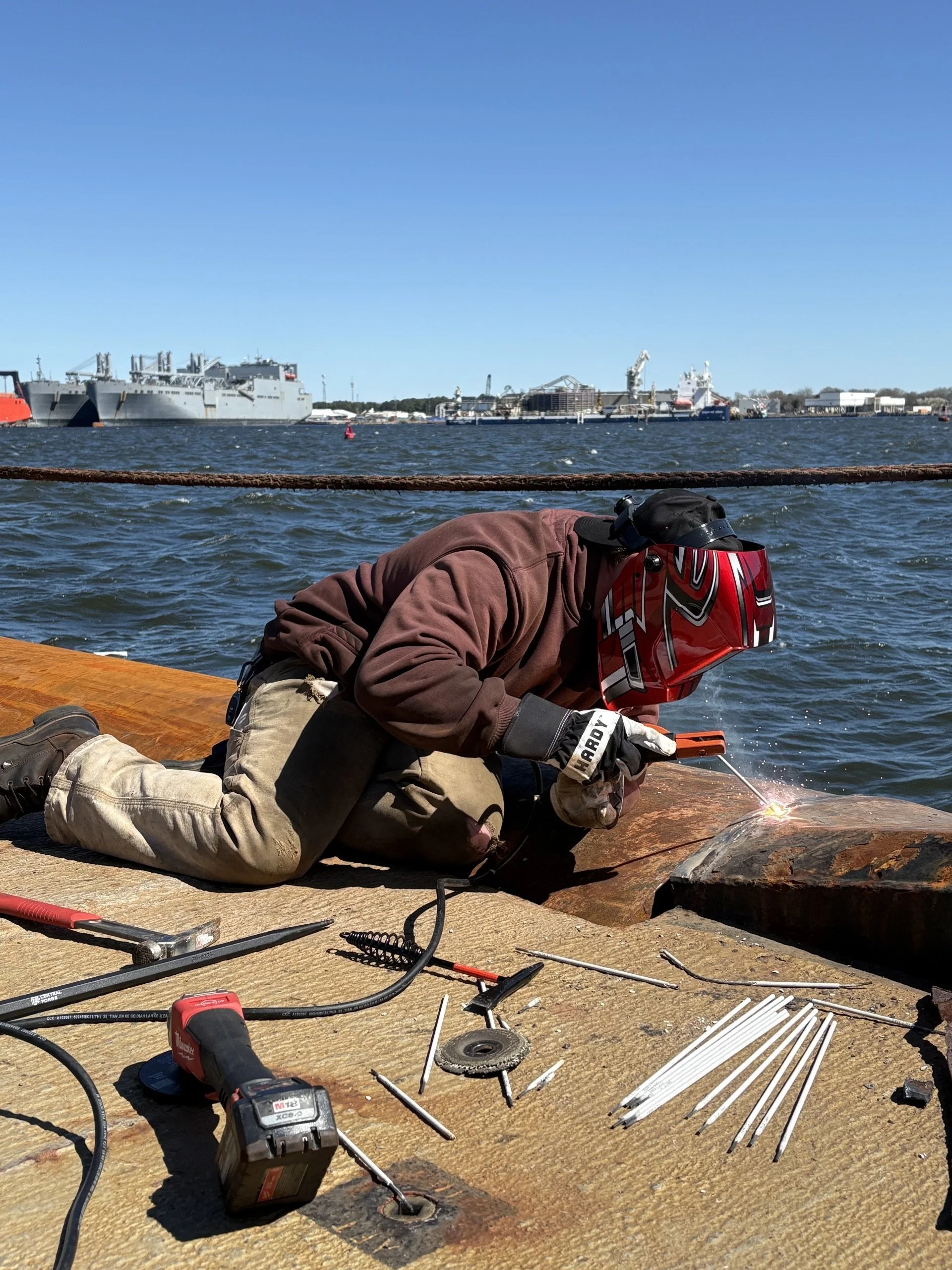 Worker welding metal on a dock by the water with ships and a port in the background.