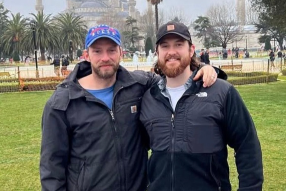 Two men standing outdoors on a grassy area, arm in arm, smiling at the camera with a park background that includes trees, a fountain, and some people.