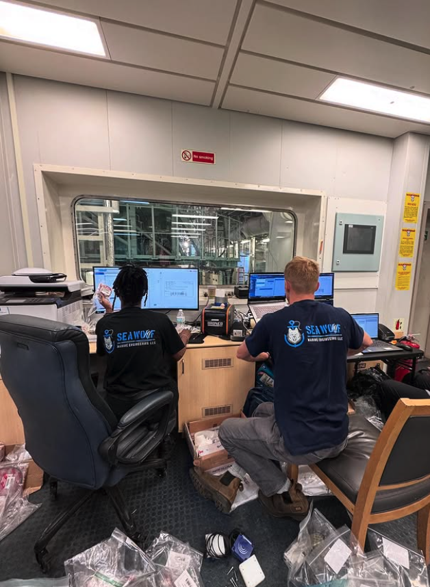 Two workers in SeaWOLF Marine Engineering LLC shirts working at a desk with multiple computer monitors in an industrial environment, with scattered supplies on the floor.