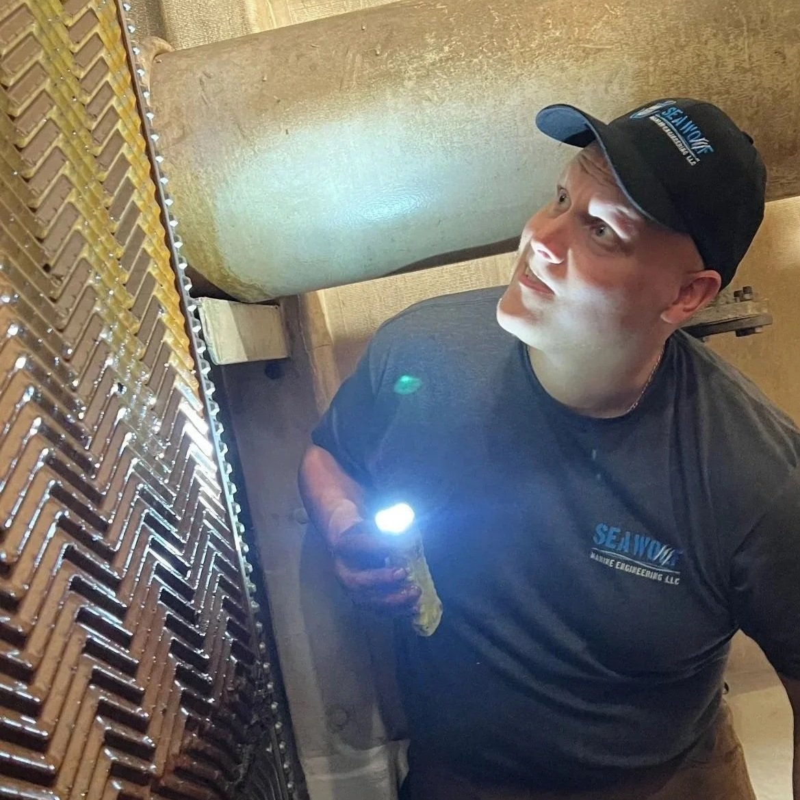 A man wearing a black cap and dark shirt holding a flashlight and inspecting a metallic, copper-colored rectangular grill with a zigzag pattern