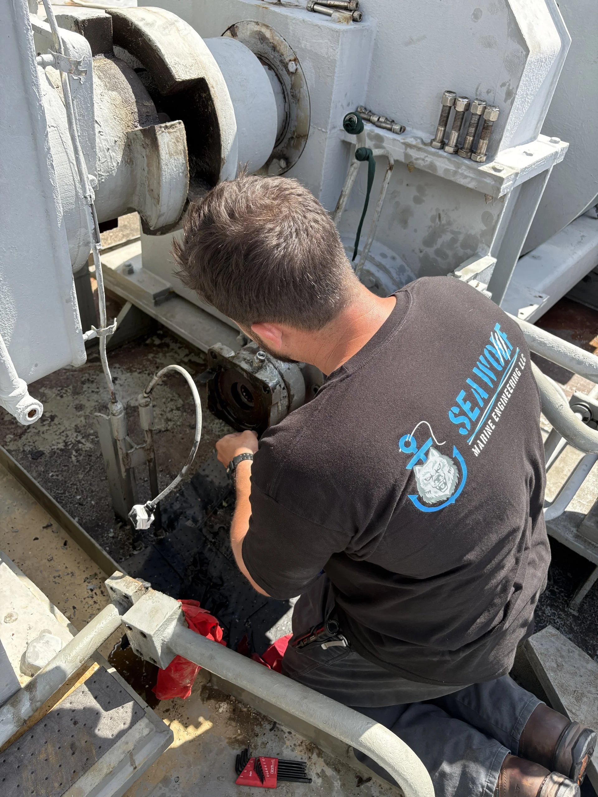 A man wearing a black Sea 101 Marine Engineering LLC t-shirt repairing or inspecting a large industrial piece of equipment outdoors, with tools and machinery parts around him.