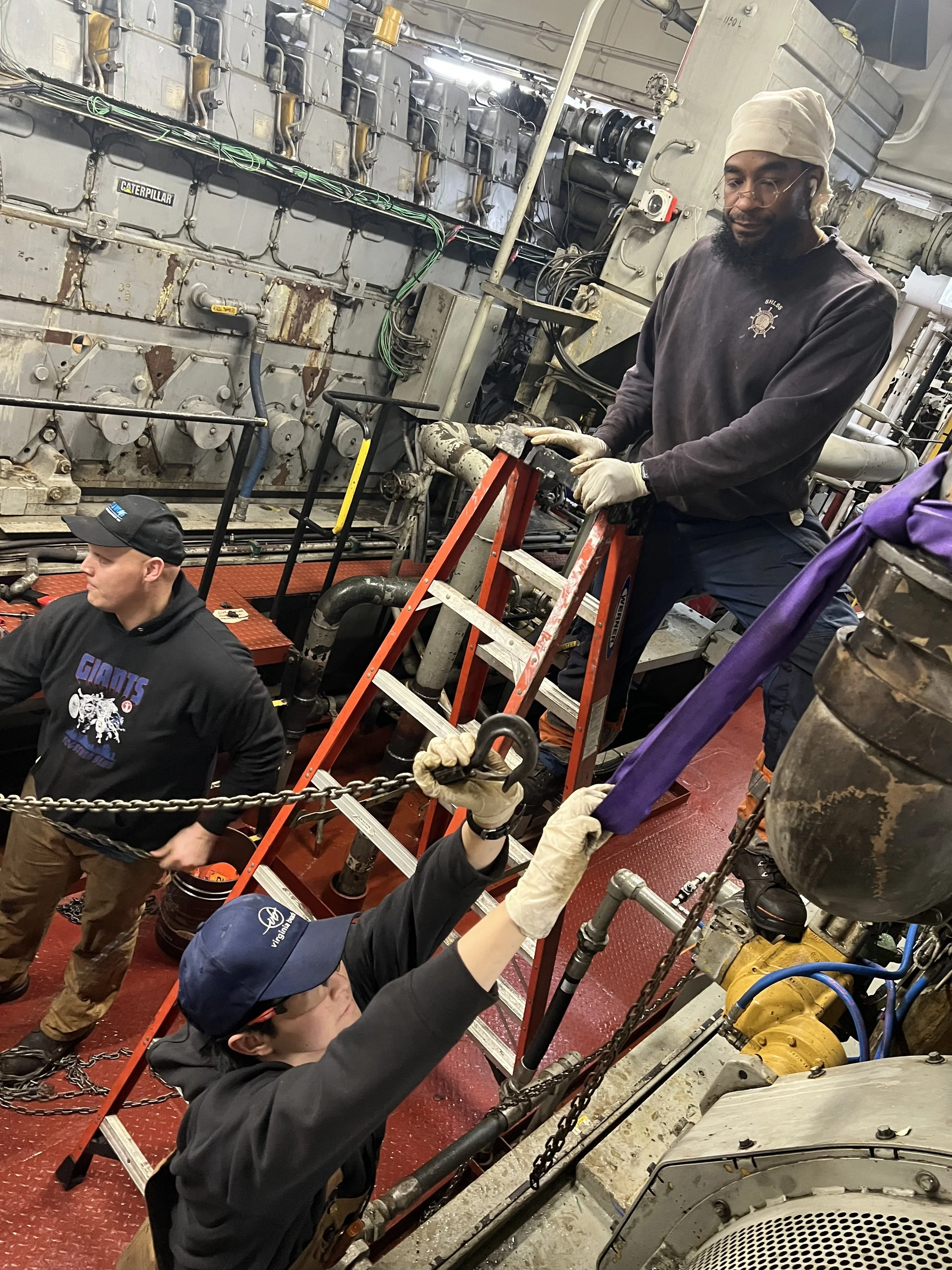 Three workers are inside an industrial engine room. One worker is on a ladder, operating a tool on a large machine. Another worker in a black shirt and cap is standing nearby, and a third worker in a cap and glasses is handing tools or equipment to the worker on the ladder. The room has large machinery, pipes, and chains.