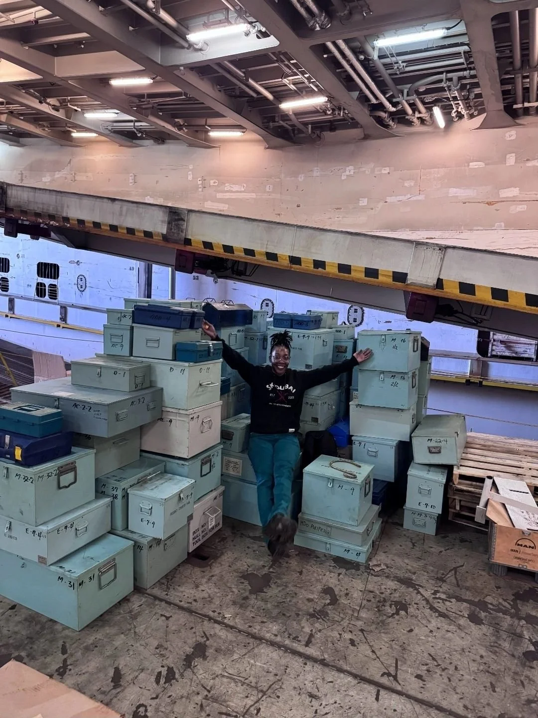 A woman standing amidst stacks of light blue storage boxes in an industrial or warehouse setting, smiling and posing with arms outstretched.