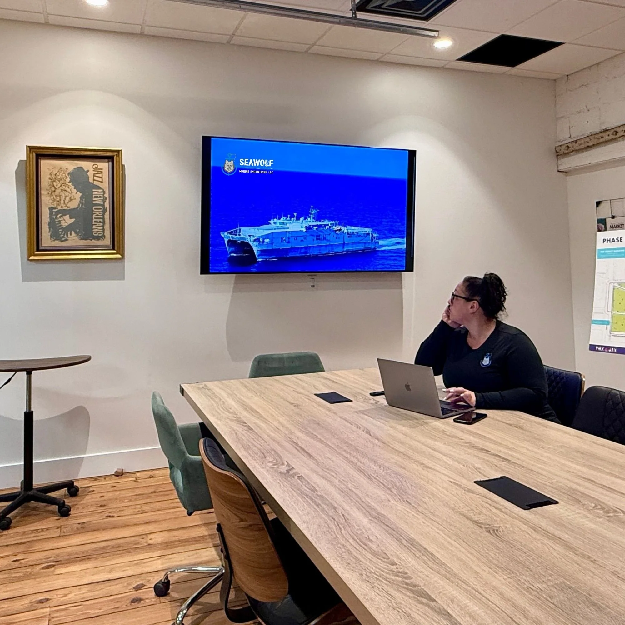 A woman sitting at a conference table in a meeting room, looking at a large screen on the wall displaying a ship and the logo "Seawolf Marine Engineering LLC." The woman is using a laptop and has a notepad and phone in front of her. There are chairs around the table, a framed picture on the wall, and a blueprint or poster on a stand partly visible on the right.