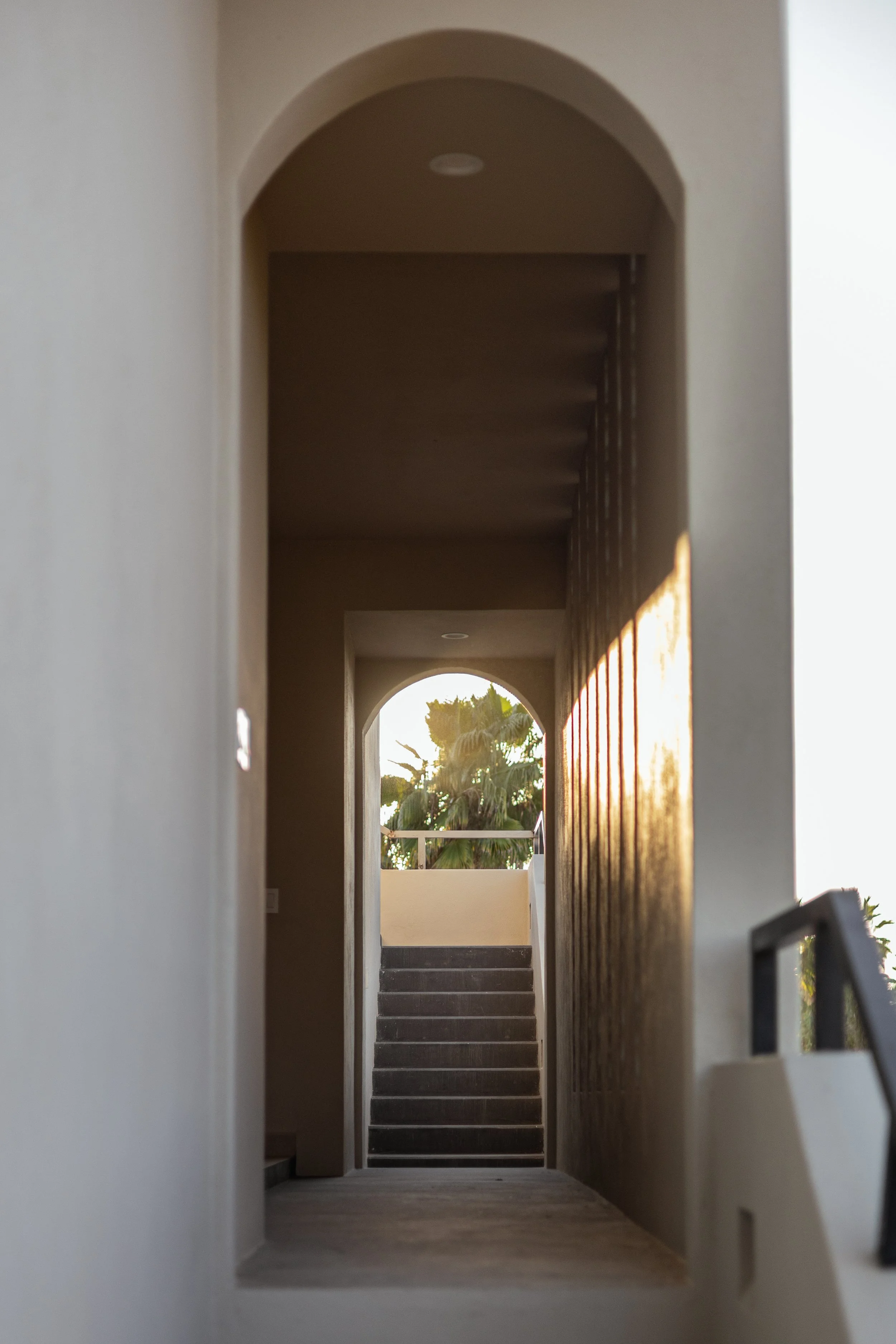 View through a narrow hallway showing stairs leading up to a balcony with palm trees in the background, sunlight casting a glow on the scene.