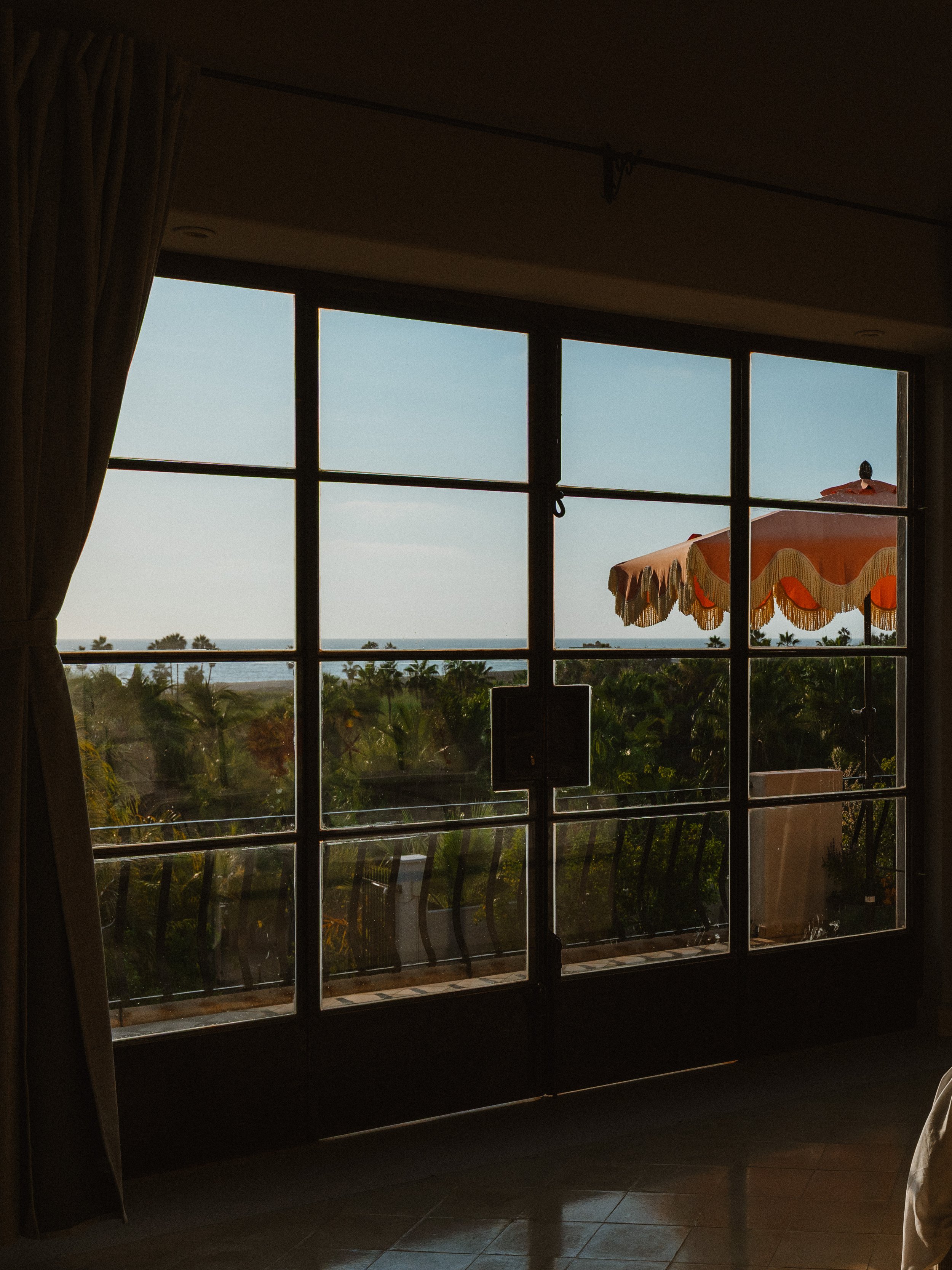 View through a glass door showing a balcony with an orange and cream decorative umbrella, lush green trees, and a distant body of water under a clear blue sky.