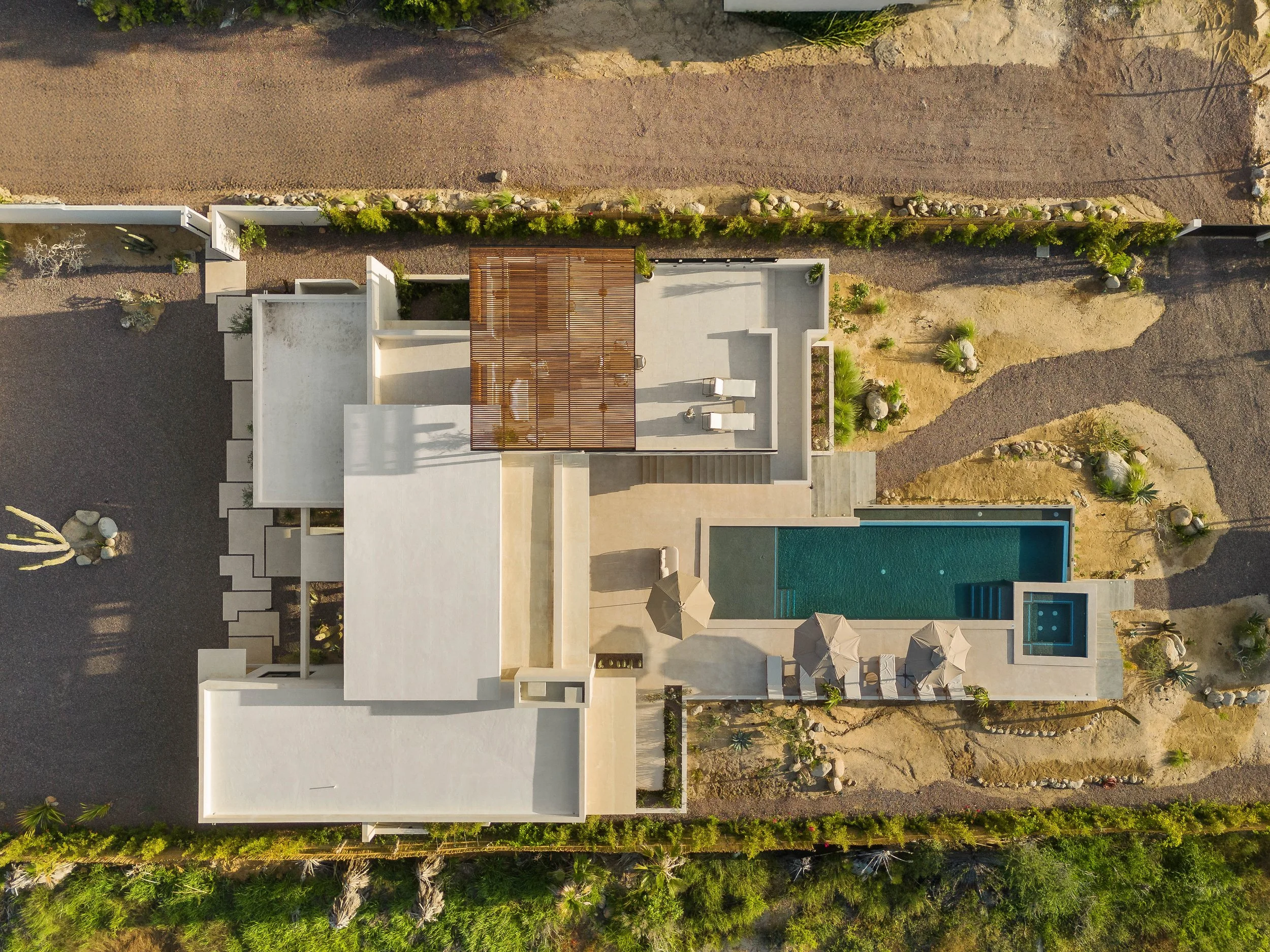 Aerial view of a modern house with a swimming pool, patio umbrellas, and surrounding desert landscape with rocks and plants.