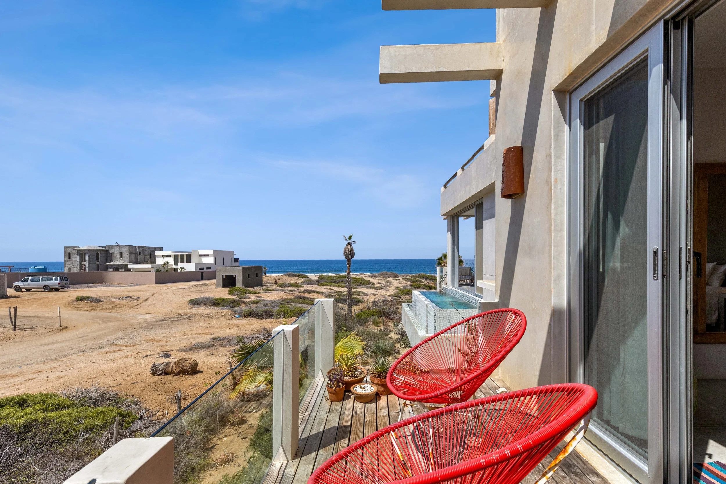 Balcony with two red lounge chairs overlooking a beach with sand, a palm tree, and ocean in the background.