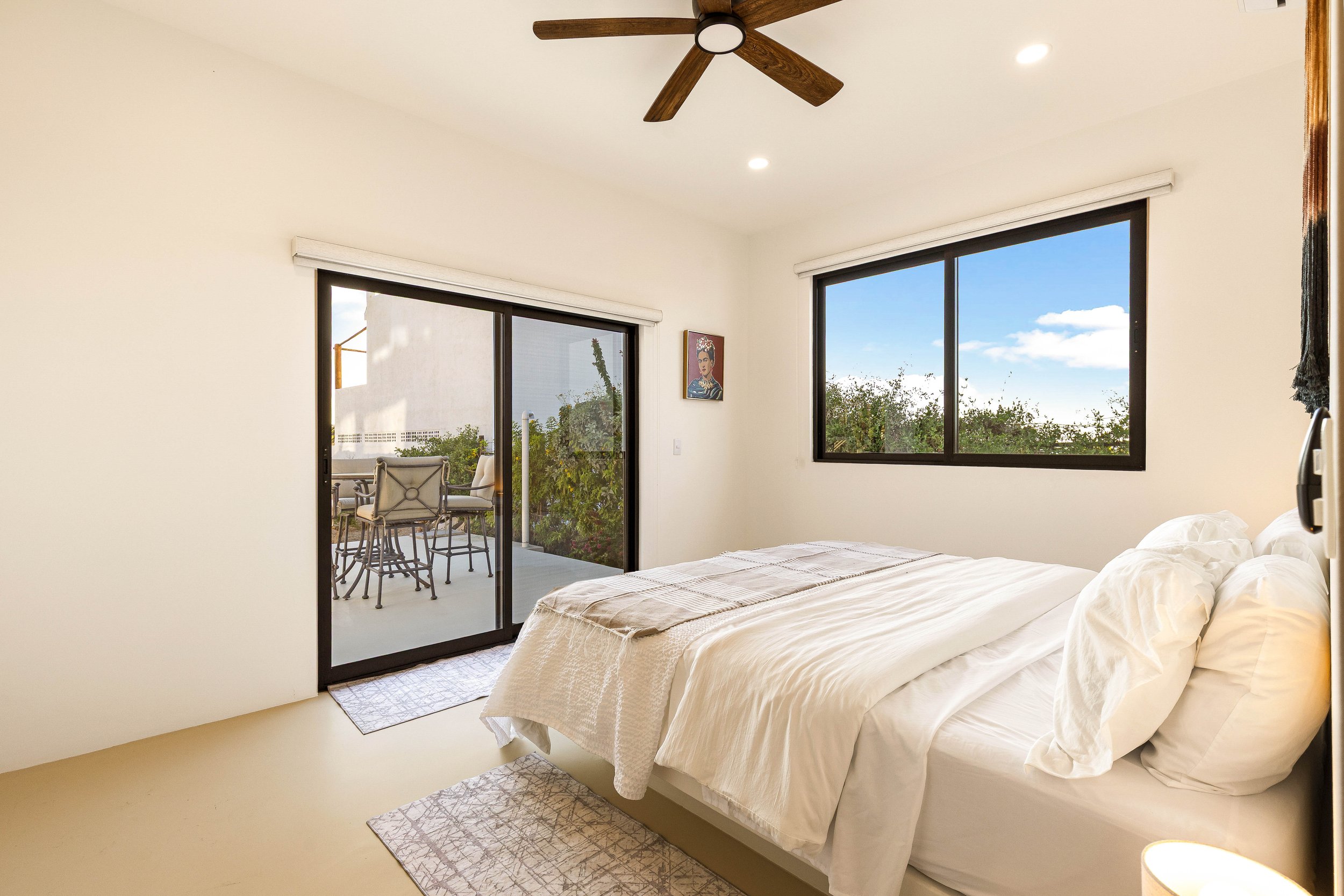 Bedroom with white bedding, a sliding glass door leading to a balcony with outdoor seating, and a window showing a blue sky with clouds.