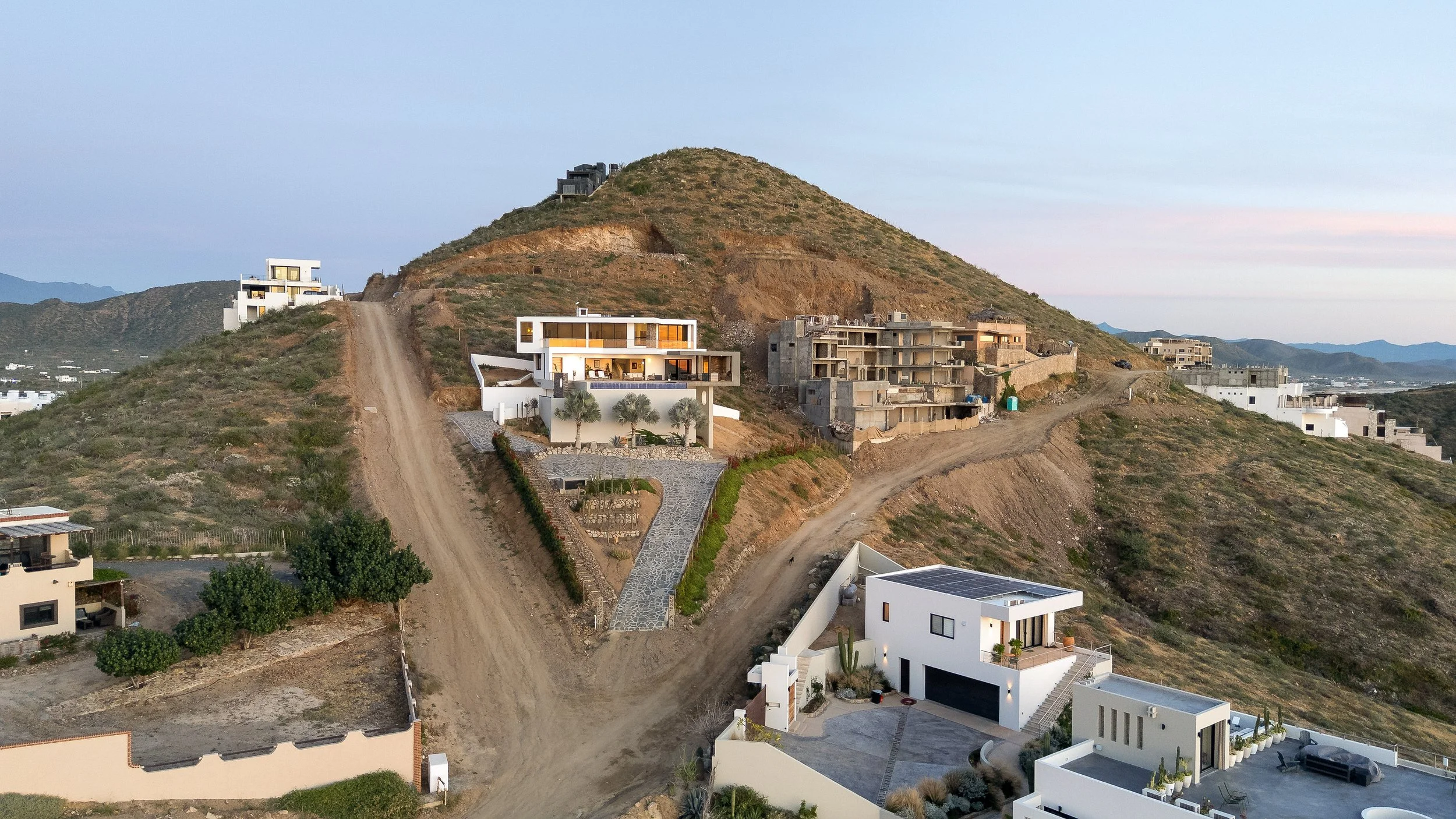 Hilly landscape with several modern white and unfinished buildings, dirt roads, and sparse vegetation with mountains in the background.