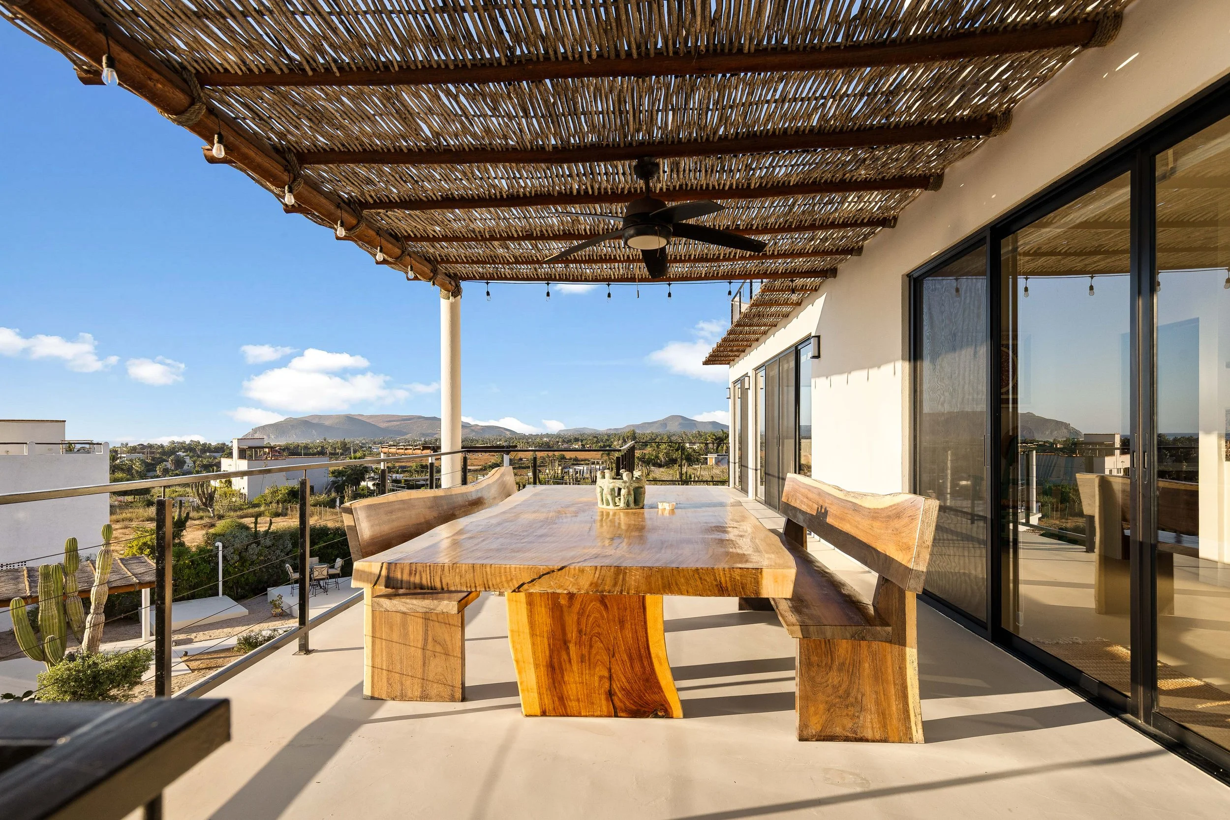 Outdoor balcony with a wooden dining table and benches, overhead bamboo shading, ceiling fan, railings, sliding glass doors, and a view of mountains and sky in the distance.