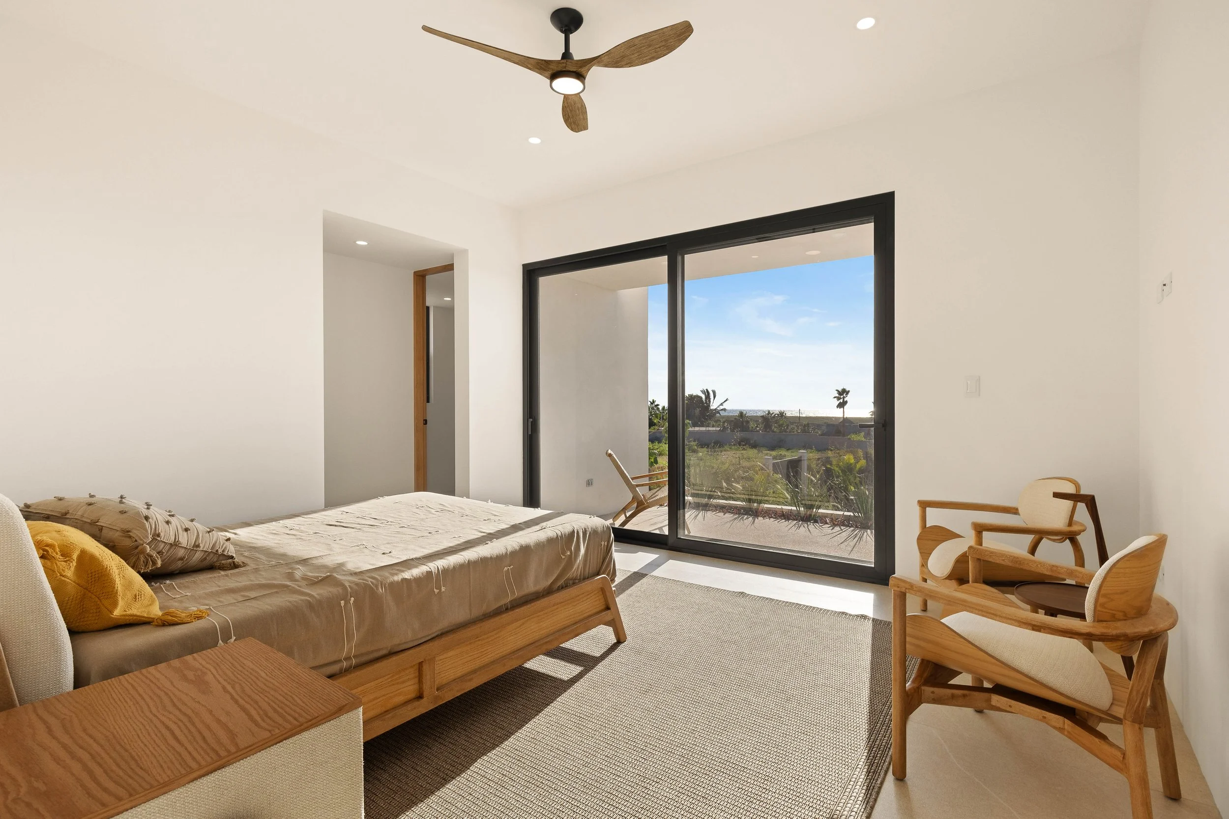 Bedroom with a sliding glass door leading to a balcony with outdoor chairs, a wooden ceiling fan, and modern wooden furniture, including a bed and two chairs, with a coastal view outside.