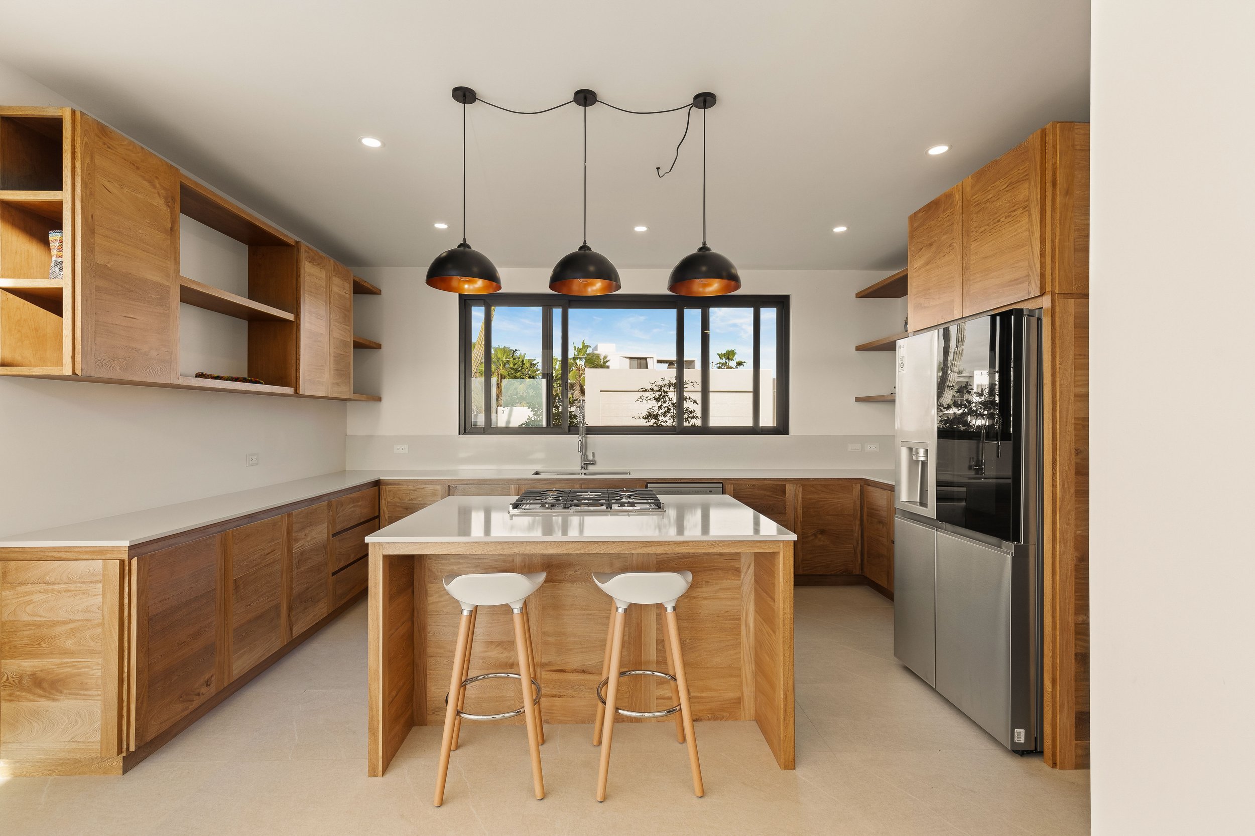 Modern kitchen with wooden cabinets and a white island with two stools, three black pendant lights, stainless steel refrigerator, black window, and a view outside.