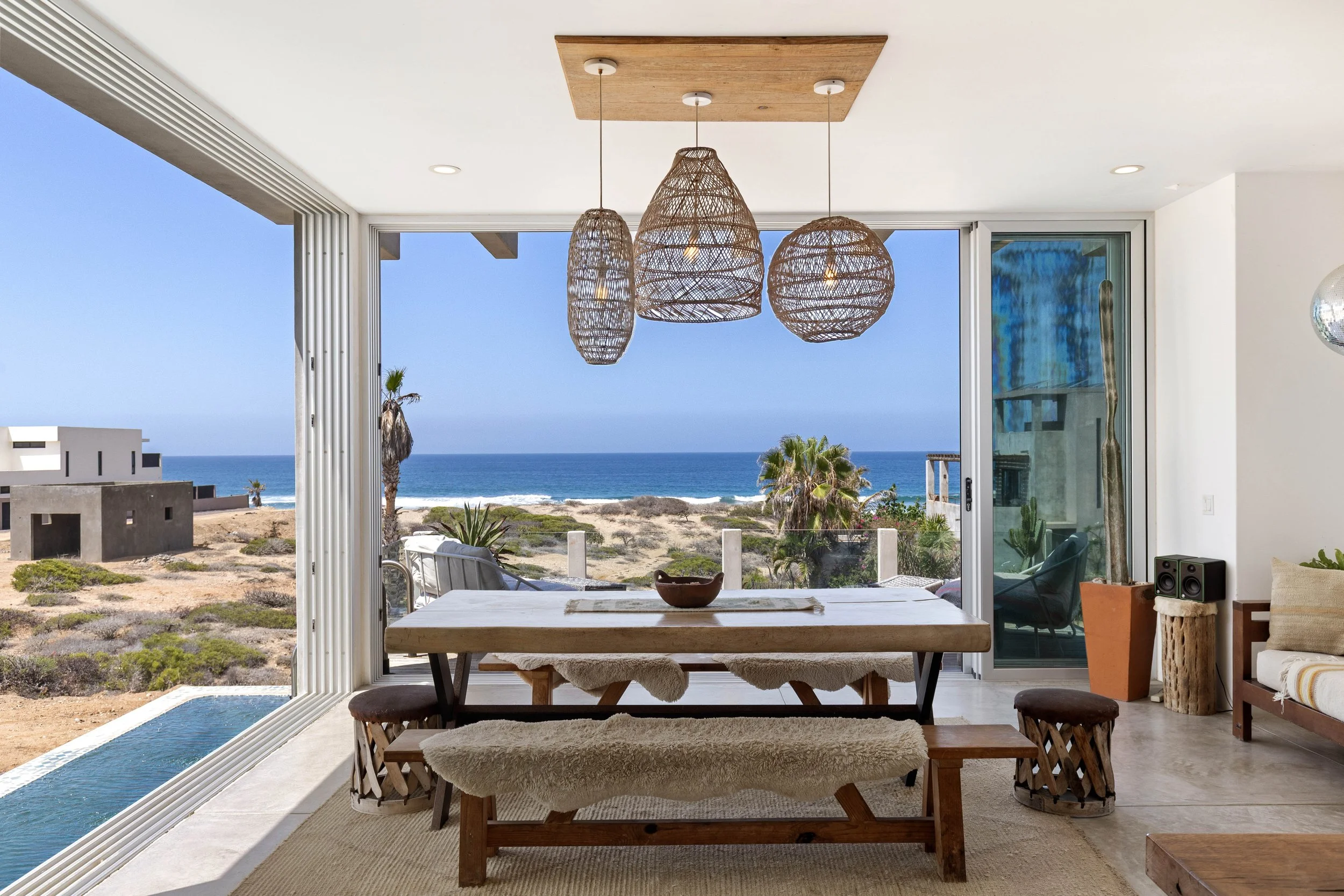 Dining area with a large wooden table and bench, overlooking a beach and ocean through large sliding glass doors, decorated with woven pendant lights and tropical plants.