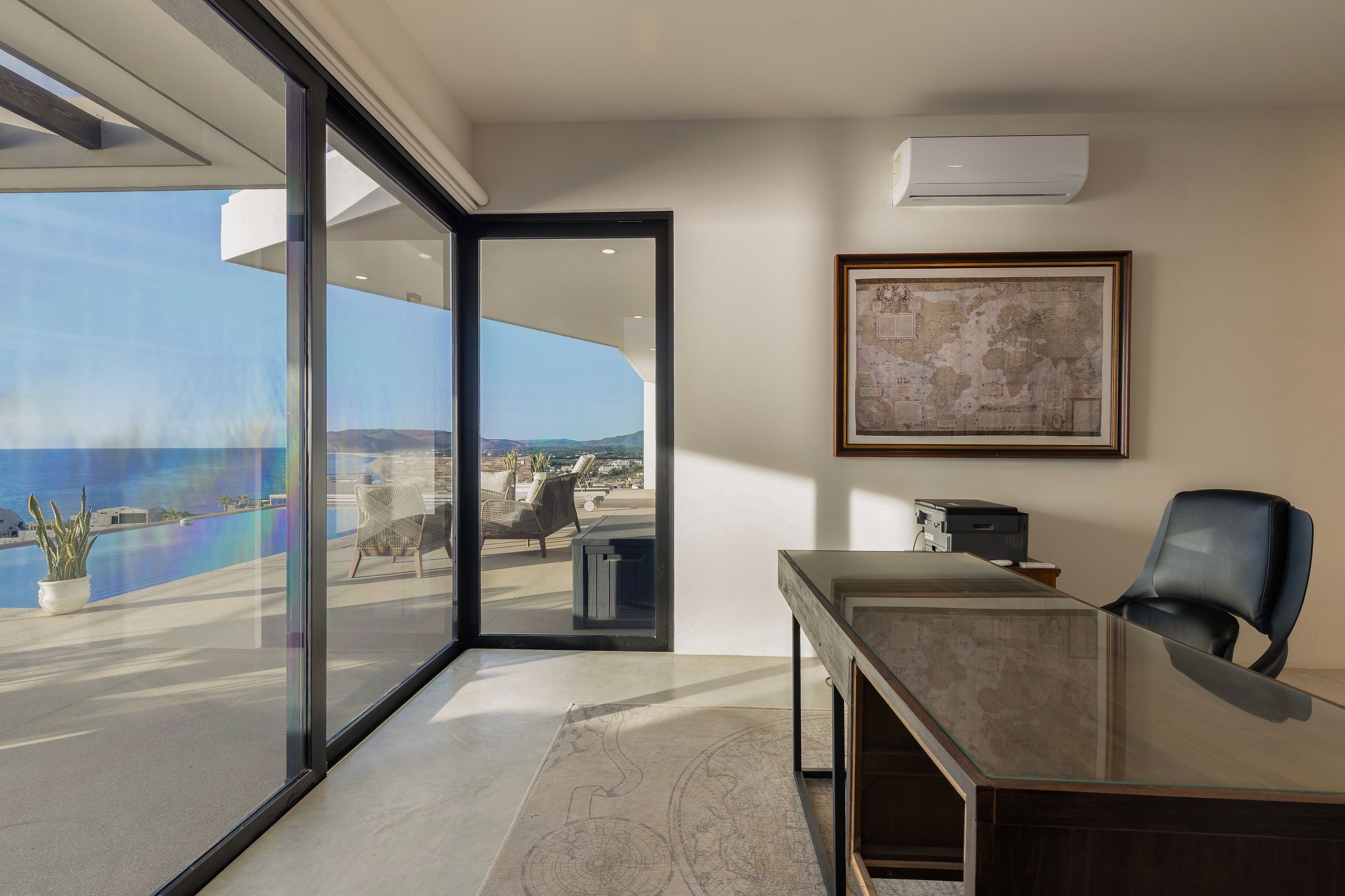 Office with large windows overlooking patio and ocean, framed world map, black office chair, wooden desk, printer, wall-mounted air conditioner.