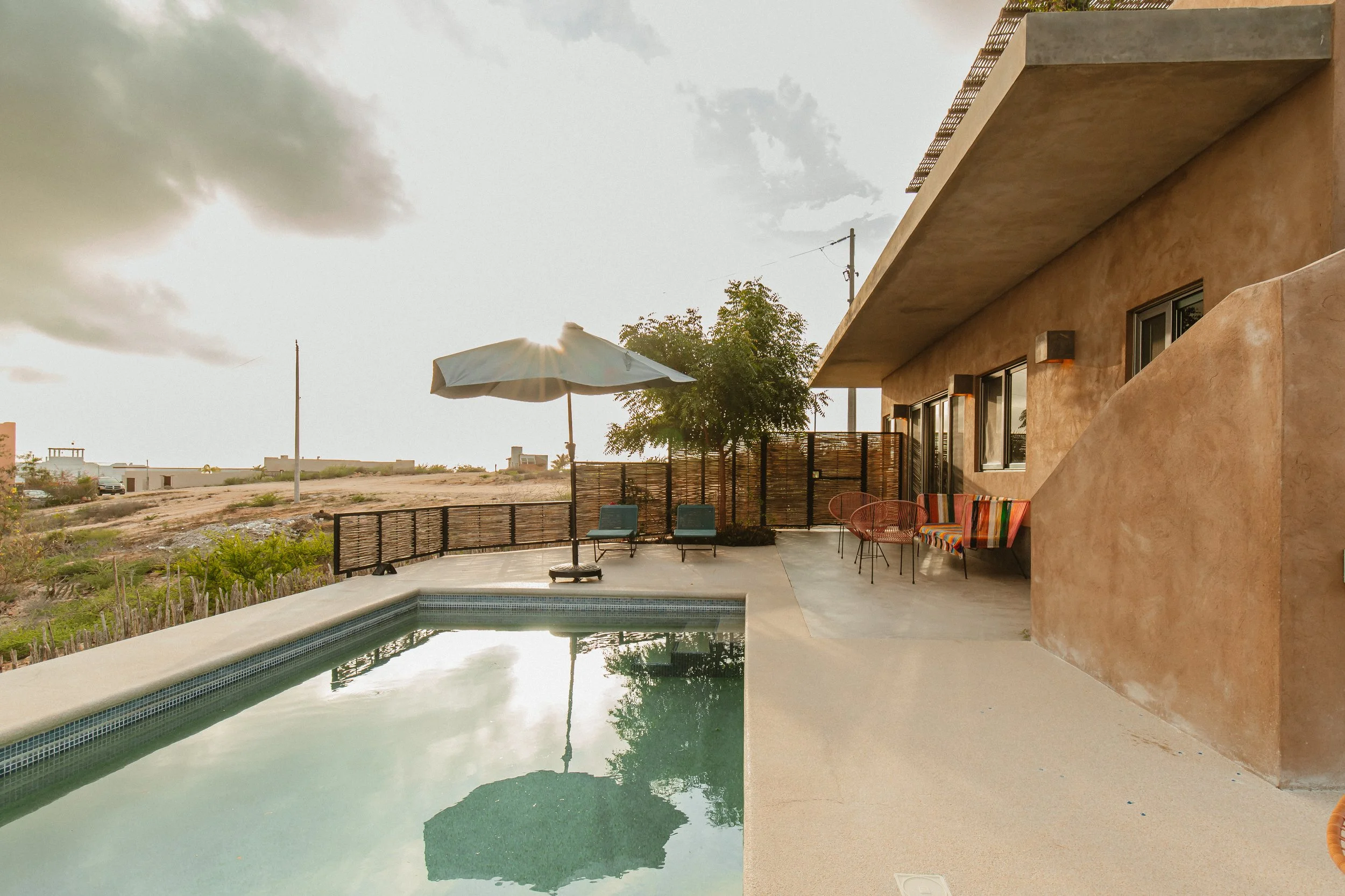 A house with a concrete patio and a small swimming pool, surrounded by a wooden fence, with outdoor furniture including chairs, loungers, and an umbrella, overlooking a desert landscape.