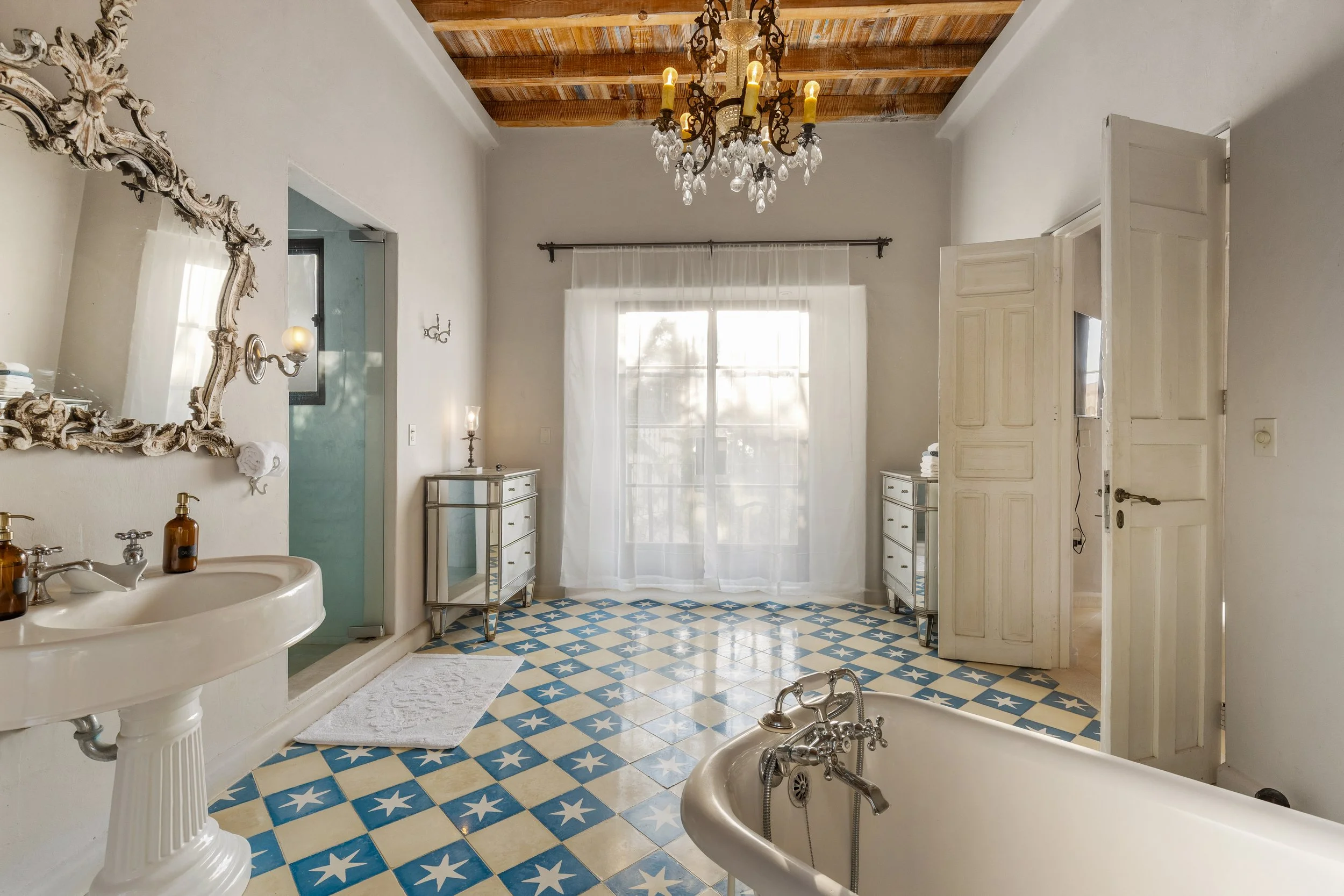 Bathroom with vintage style featuring a clawfoot tub, a pedestal sink, patterned blue and white tiled floor, a chandelier, a large window with sheer white curtains, and two mirrored dressers.