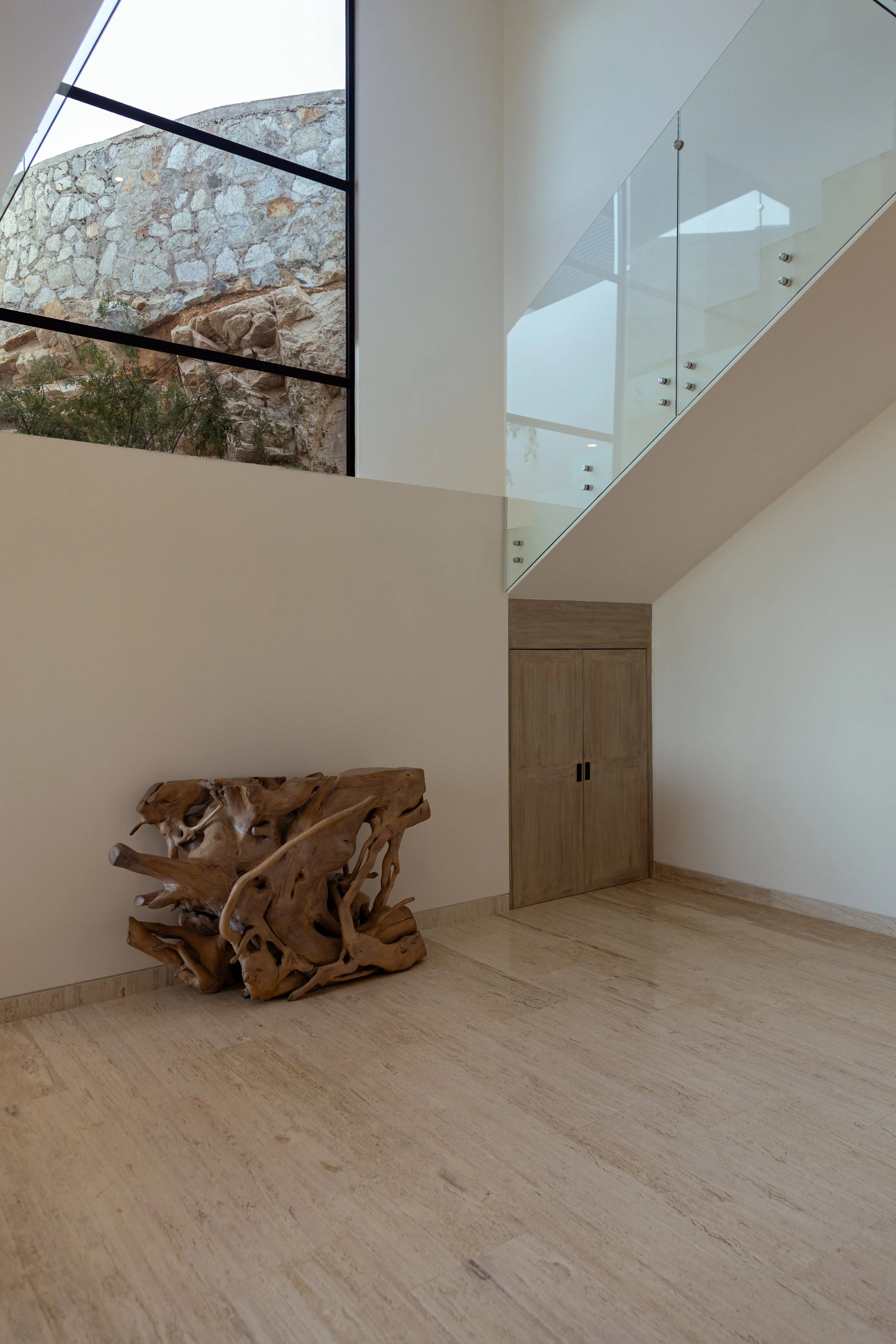 Modern interior room with a wooden sculptural art piece on the floor, light-colored wood flooring, white walls, a built-in wooden cabinet, a large window with a view of a stone wall and rocks outside, and a glass staircase with metal fasteners.