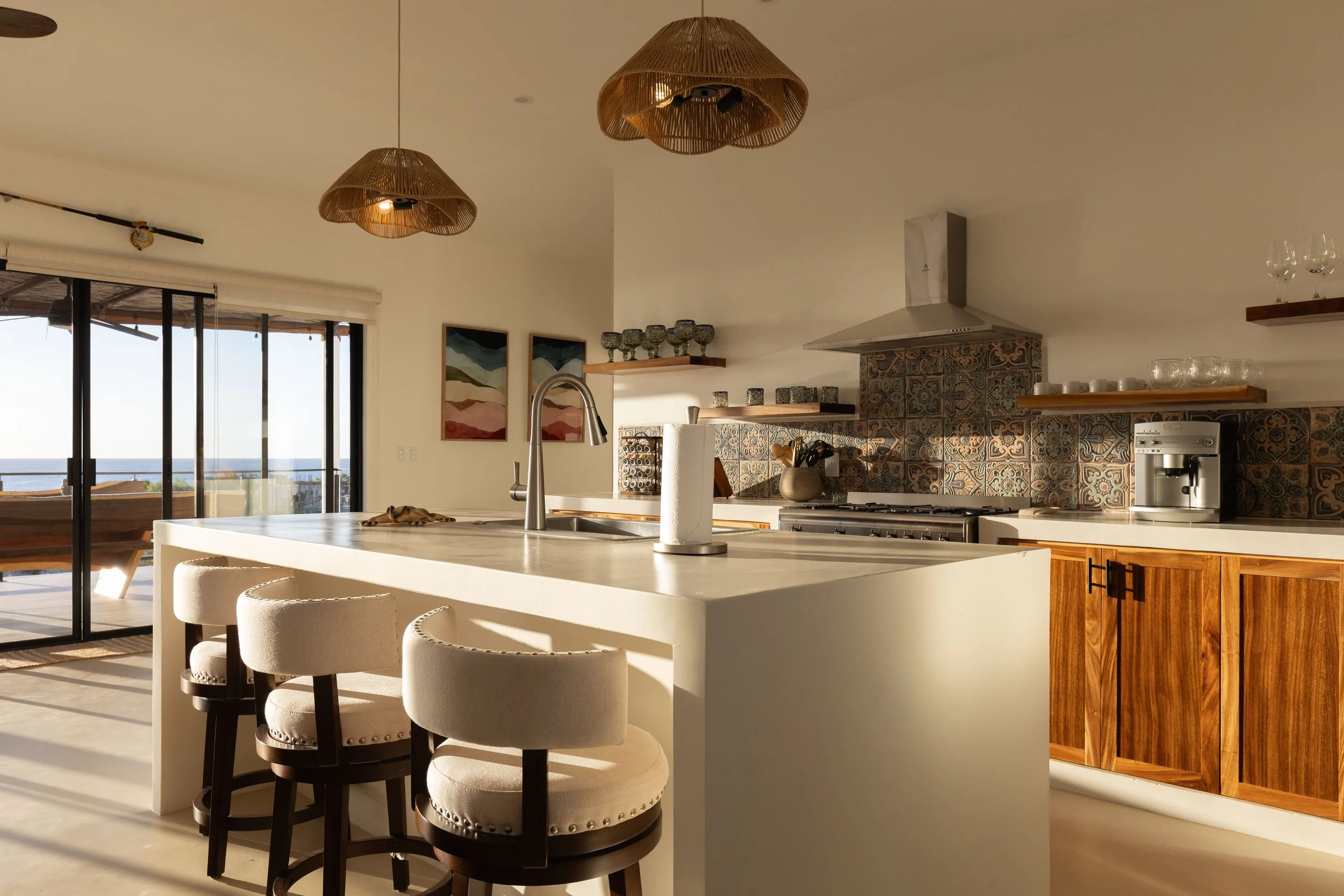 Modern kitchen with white island, three beige barstools, patterned backsplash, wooden cabinets, and sliding glass doors leading to a balcony with sea view.