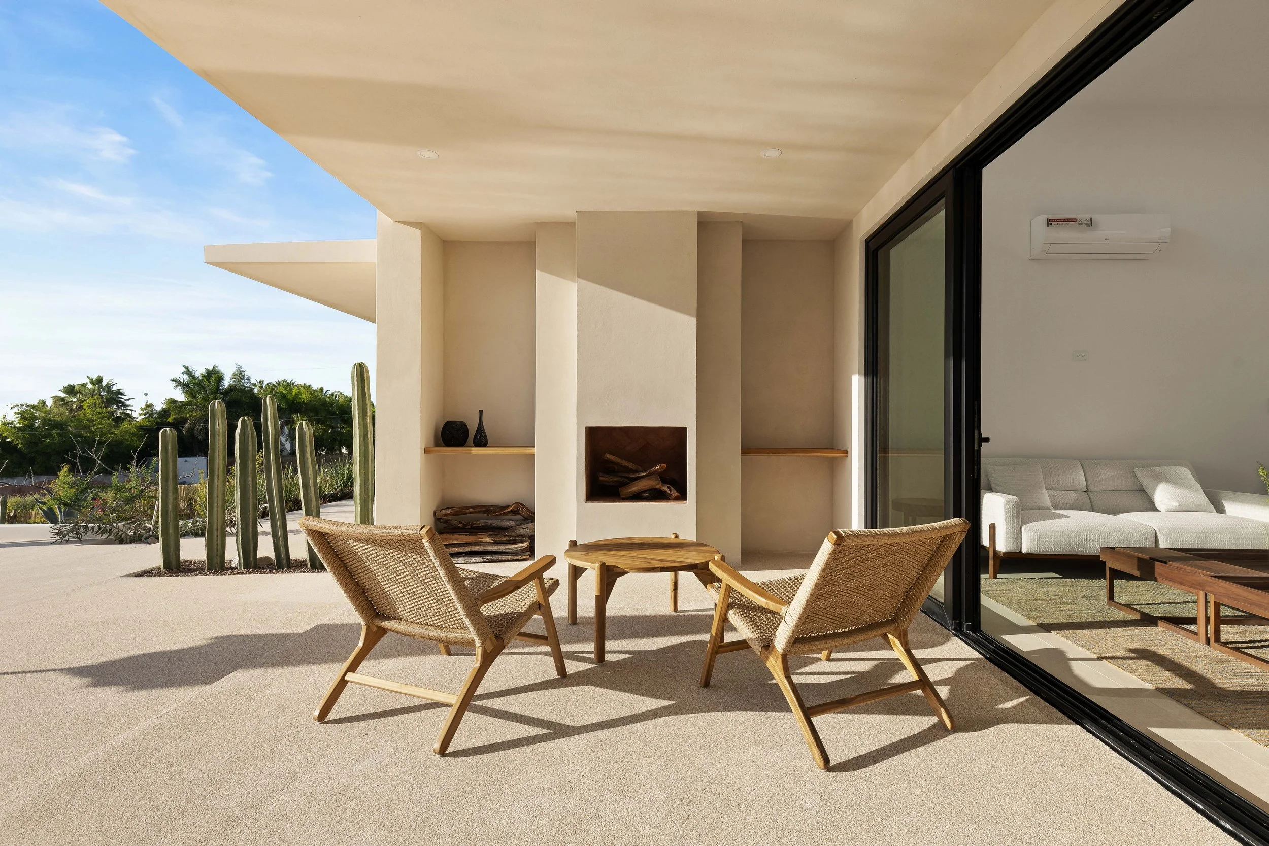 Outdoor patio with two woven armchairs and a small round wooden table, fireplace built into wall, sliding glass door leading to a living room with white sofa, desert plants and cacti outside, bright sunny day.