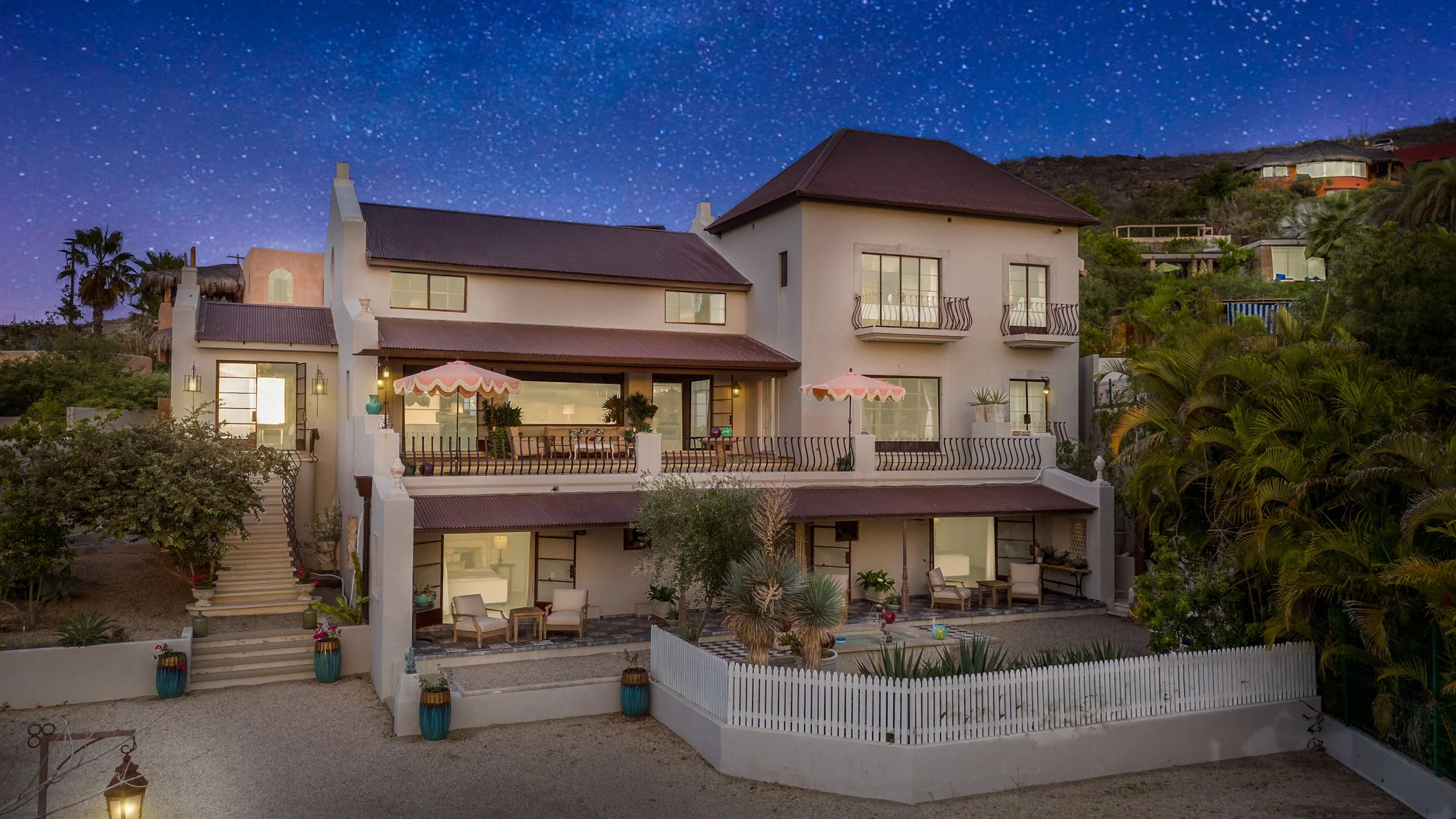 A multi-story house at night with illuminated windows and outdoor seating areas, surrounded by greenery and a starry sky.