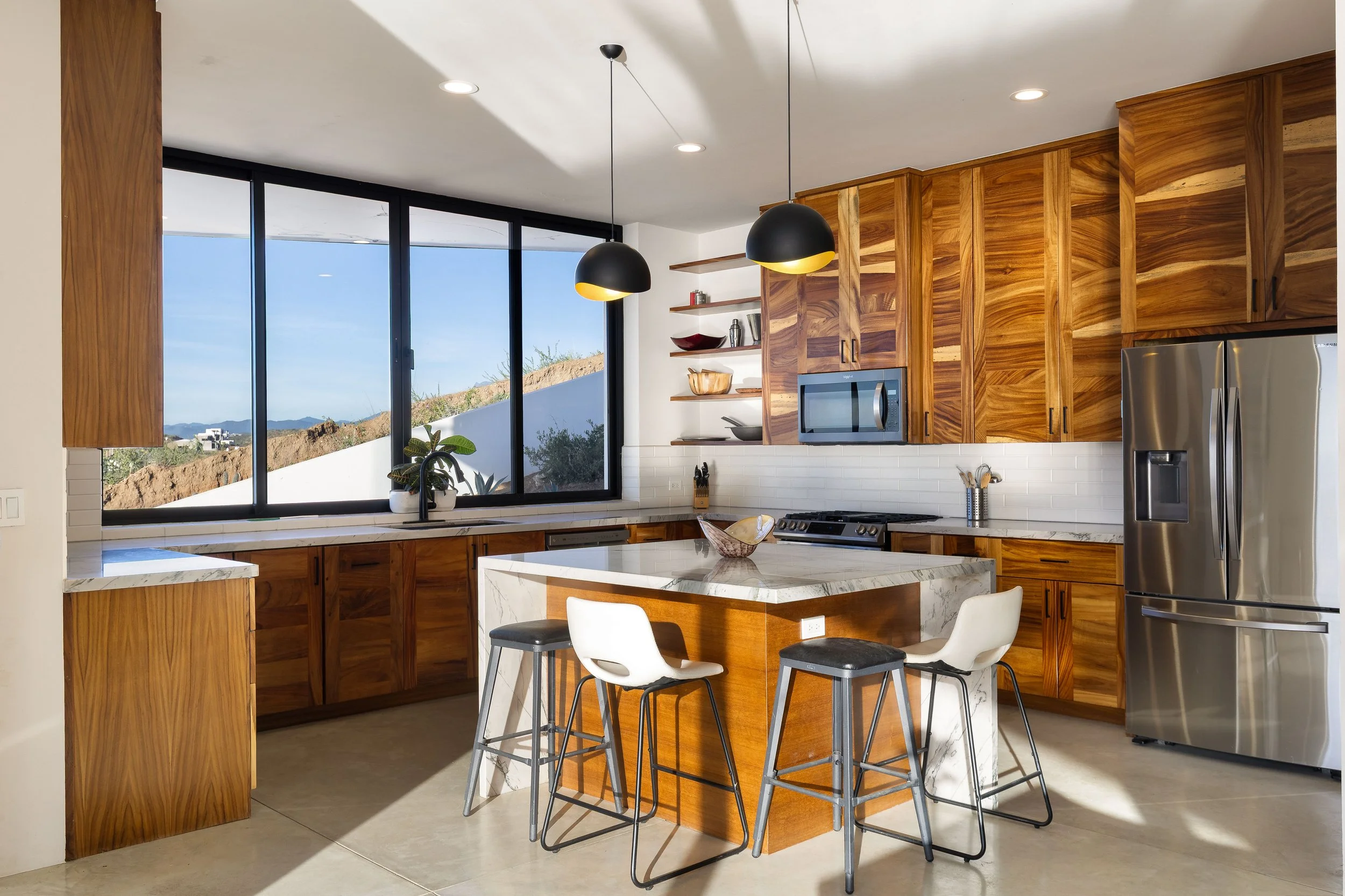 Modern kitchen with wooden cabinets, a marble island, black pendant lights, large window, white subway tile backsplash, stainless steel refrigerator, microwave, and bar stools.