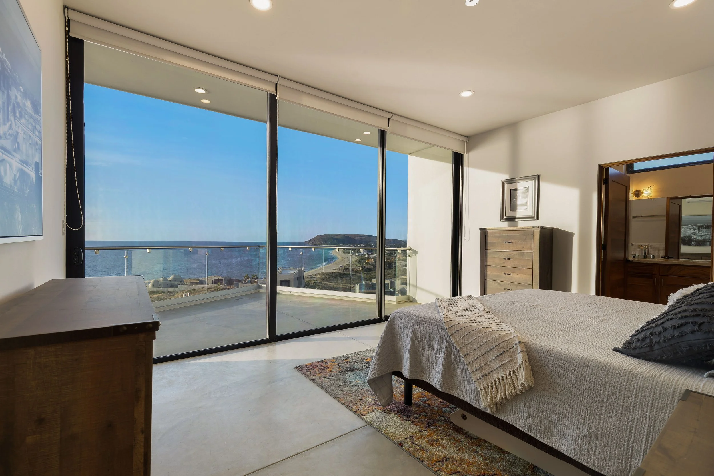 Bedroom with large glass sliding doors leading to a balcony with ocean and land view, bed with beige blanket and dark pillows, wooden dresser, and doorway to bathroom.
