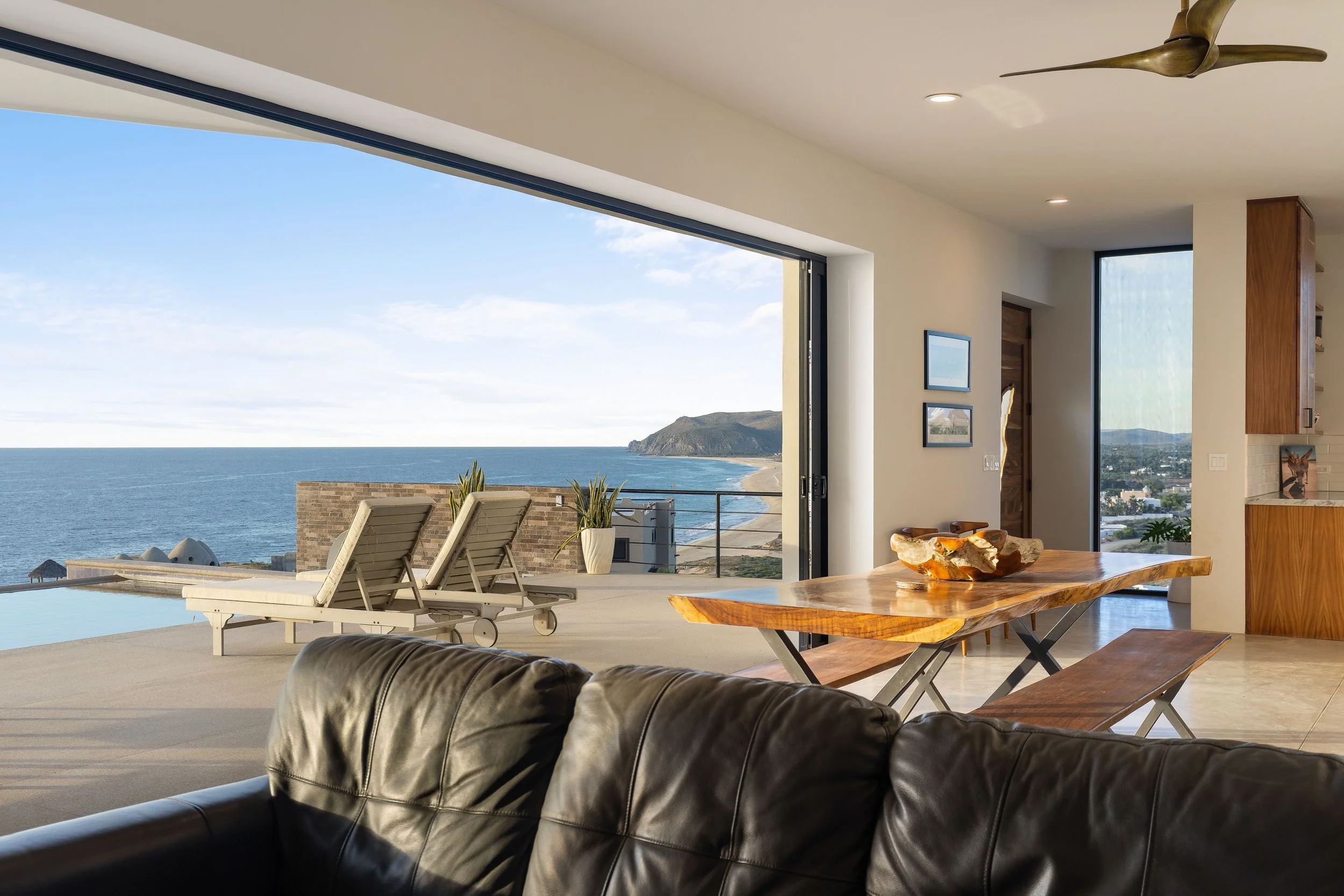 Living room with large open sliding glass door leading to outdoor balcony with ocean view, outdoor sofa, two lounge chairs, and a wooden dining table inside.