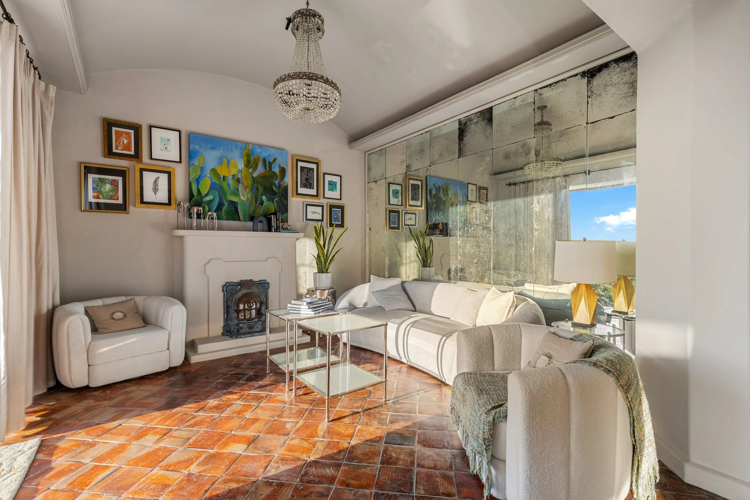 Living room with white couches, a glass coffee table, plants, a fireplace, and artwork on the wall. Large mirror on the right wall reflects a chandelier and window showing blue sky.