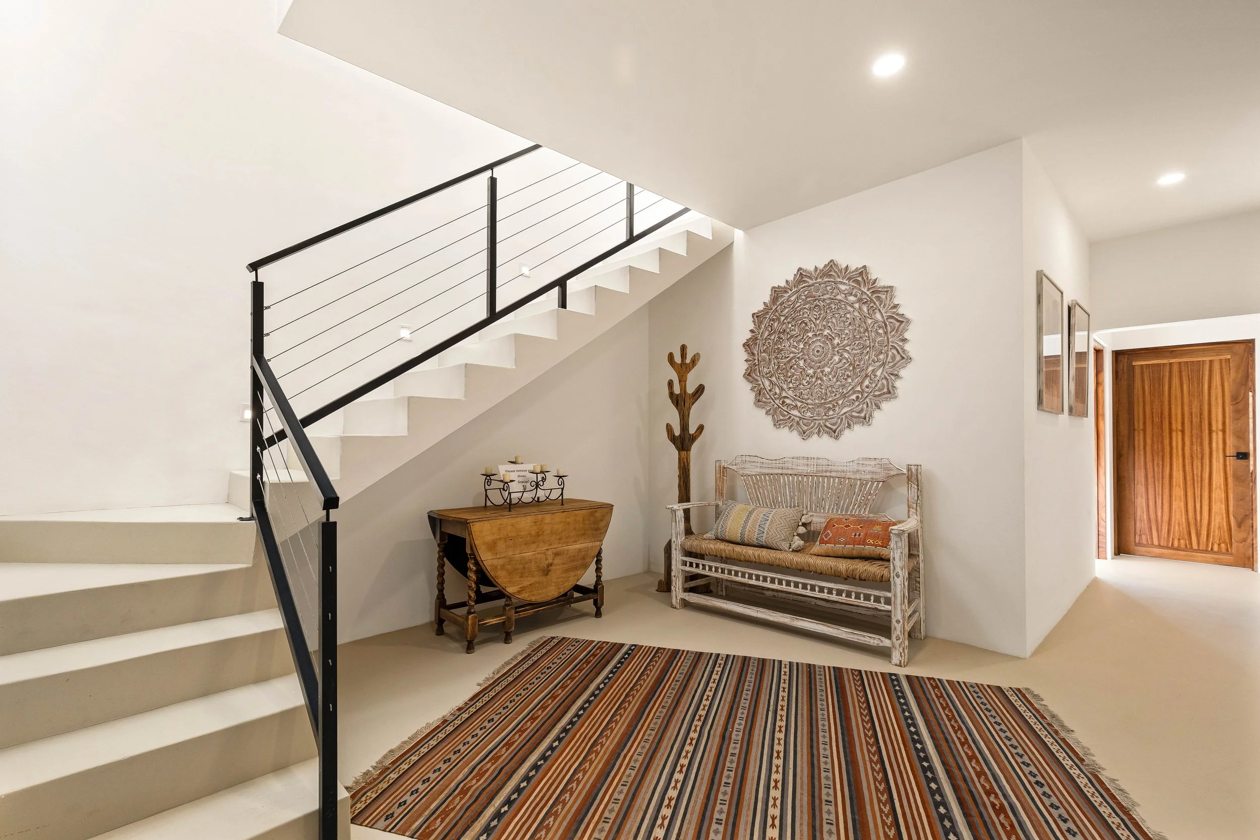Interior of a modern entryway with a staircase, decorative wall art, and a seating area with a rug, pillows, and wooden furniture.