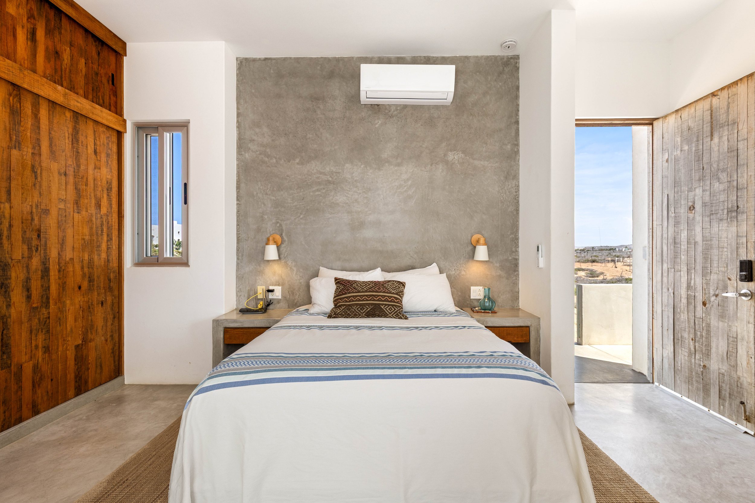 Modern bedroom with concrete and wooden walls, a bed with white and patterned pillows, bedside lamps, and an open door leading to outdoor scenery.