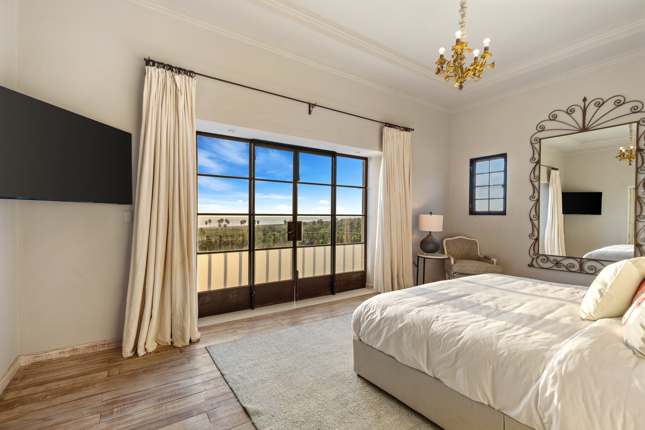 A cozy bedroom with a large bed, a bedside lamp, a wall mirror, a small window, and a sliding glass door leading to a balcony with a scenic view of trees and a blue sky.