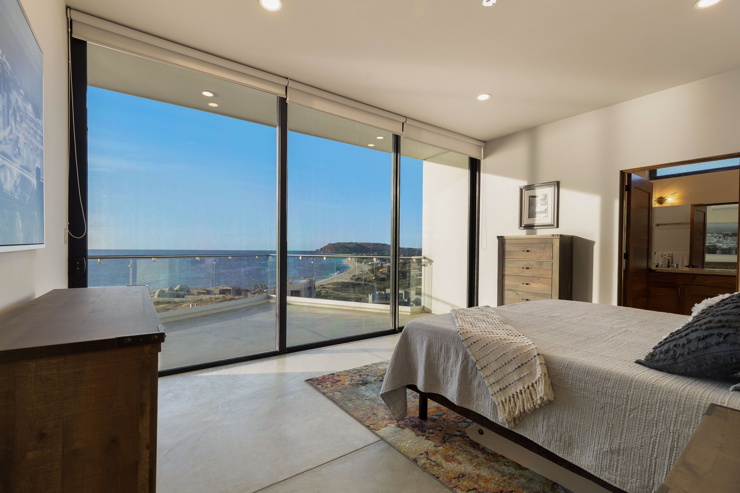 Bedroom with a large glass sliding door overlooking a coastal view, a bed with beige bedding and a blanket, a dresser, and an adjoining bathroom.