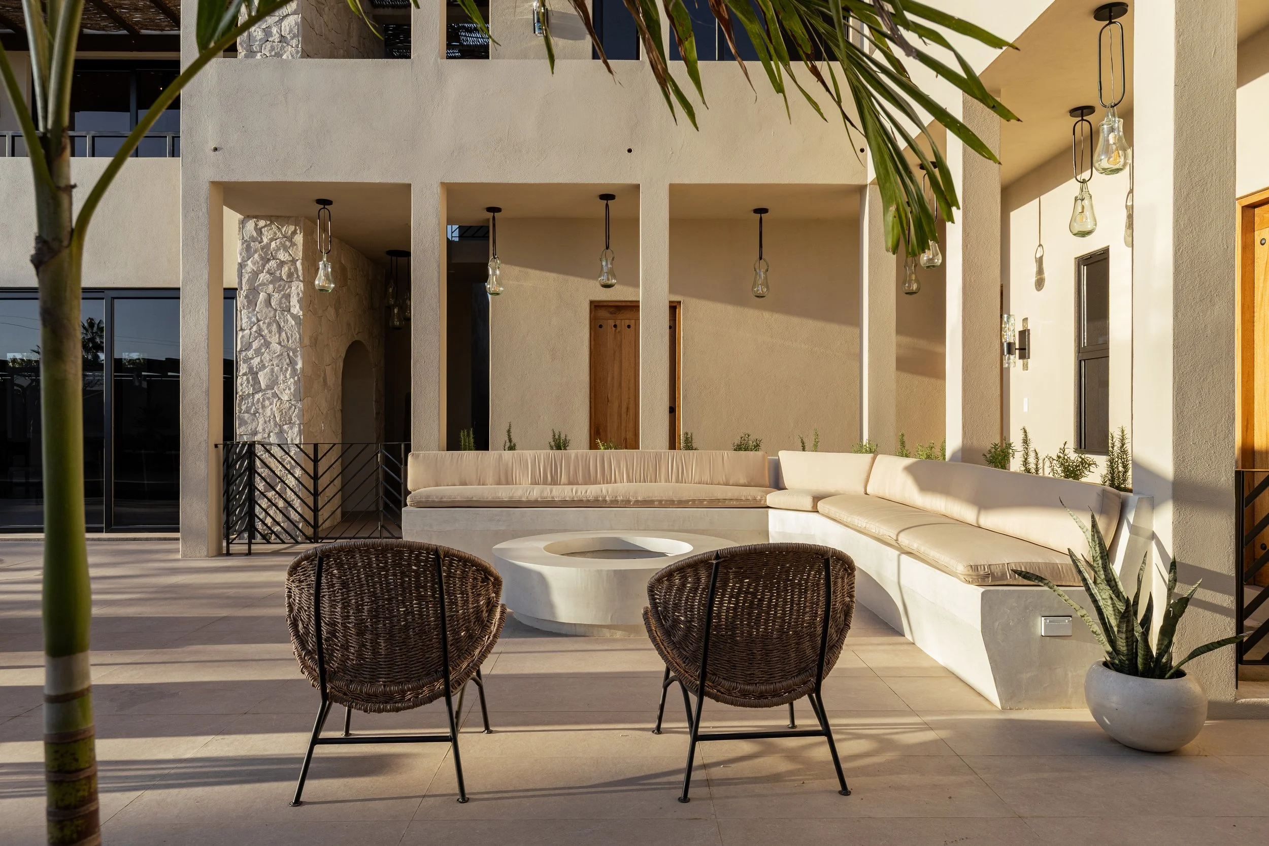 Outdoor patio with built-in white concrete bench with beige cushions, two dark wicker chairs, a white round fire pit table, potted plants, and hanging light bulbs, with a beige stucco building in the background.