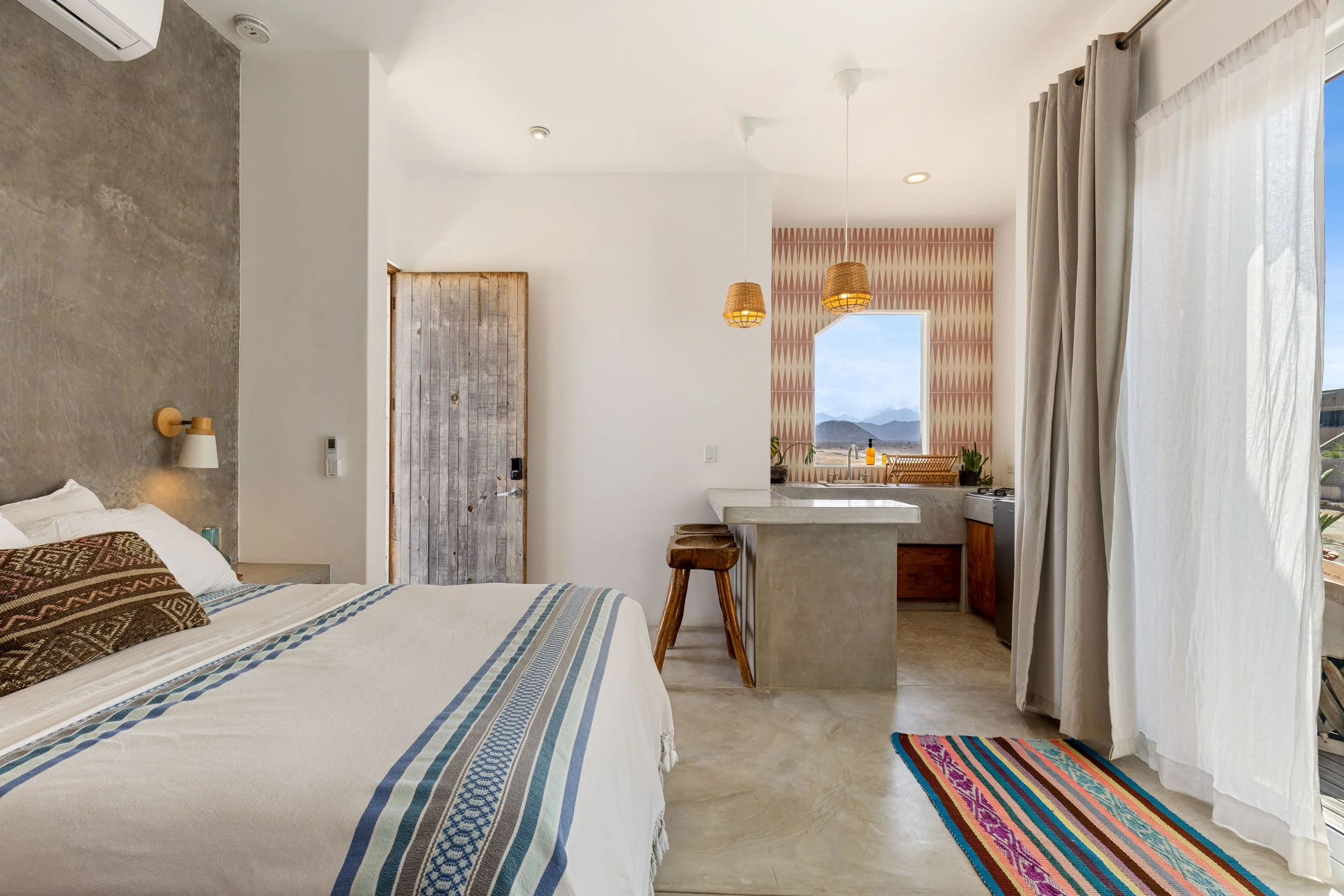 A hotel room with a bed, nightstand, wall lamp, wooden door, kitchenette with hanging lights, window with a view of mountains, patterned wallpaper, and a colorful rug near white curtains.