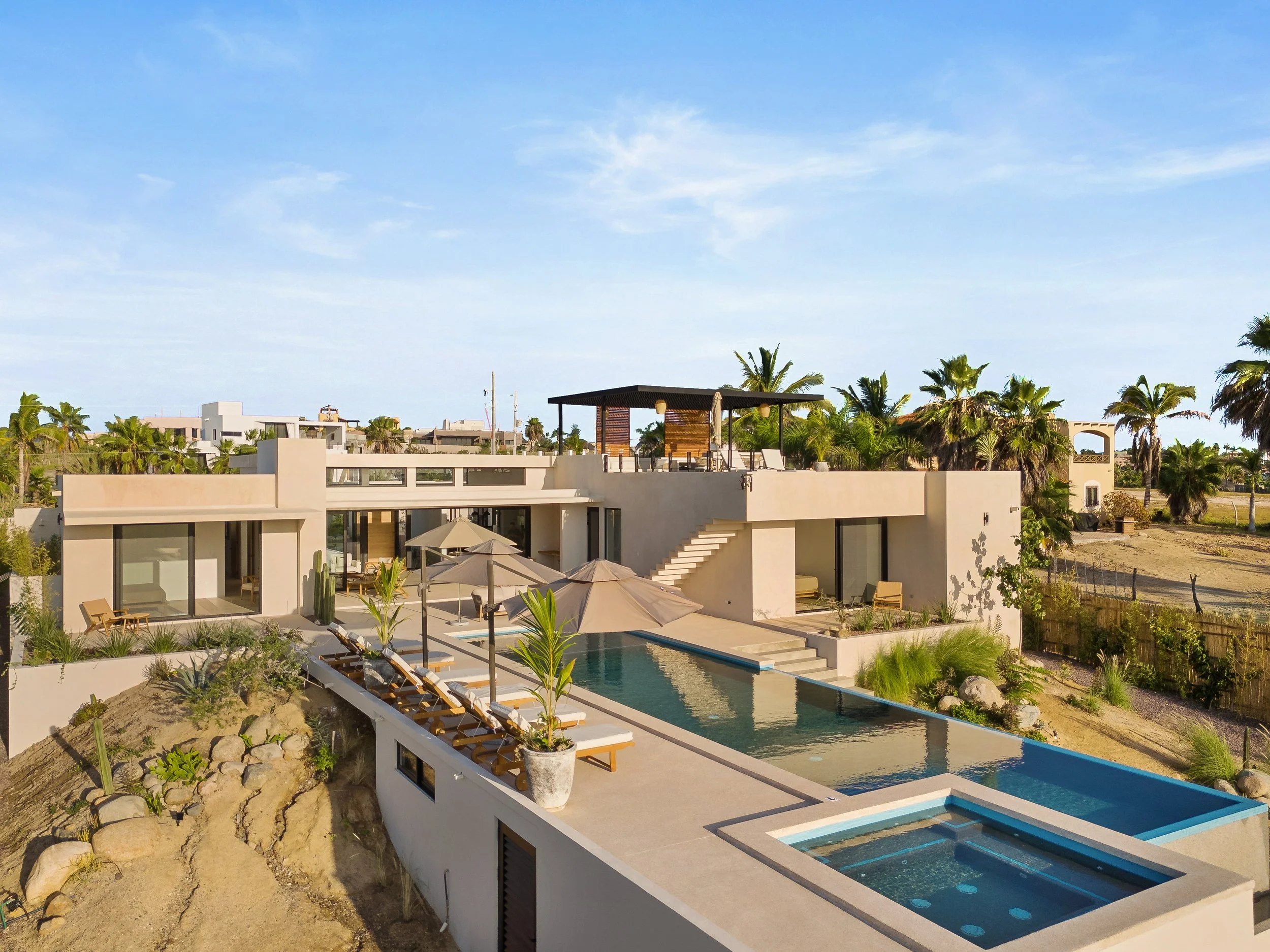 Modern house with swimming pool, outdoor lounge chairs, umbrellas, and lush palm trees.