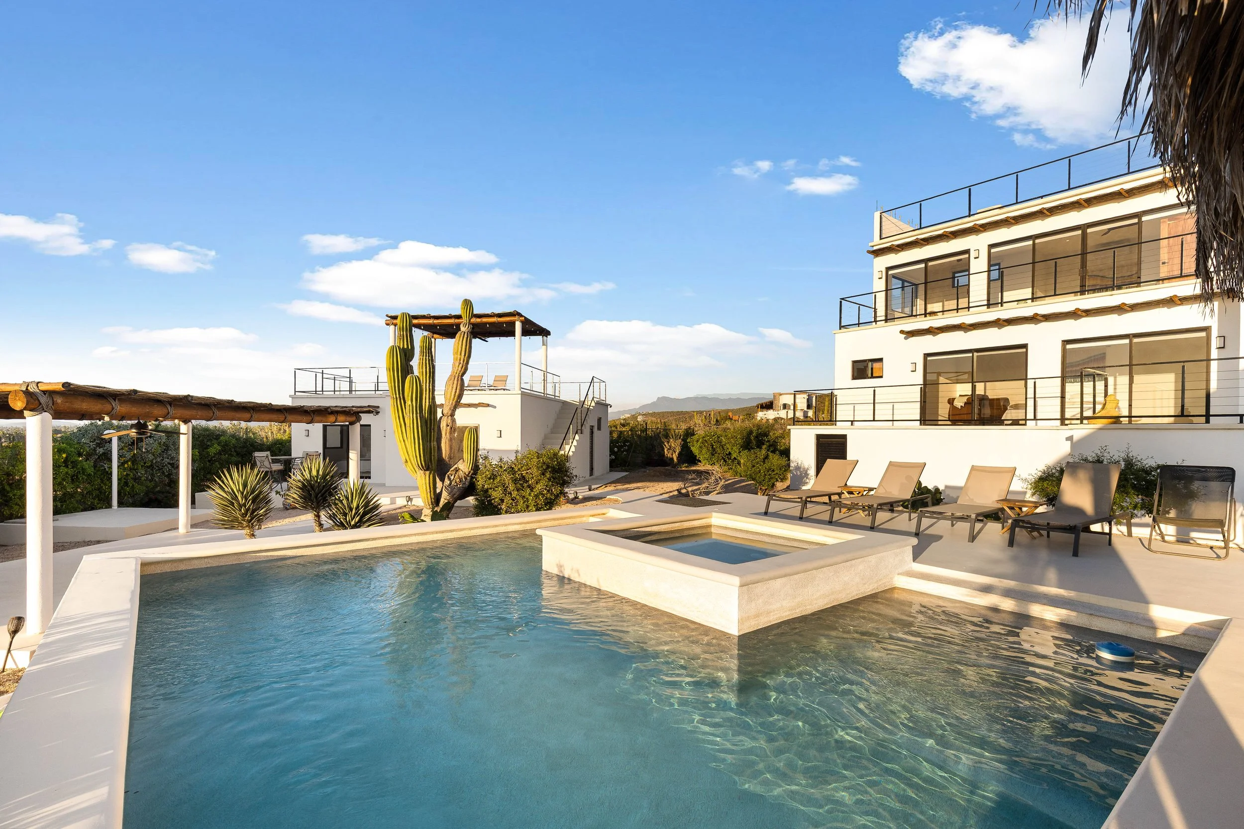 Modern white multi-story house with large glass windows and balconies, swimming pool with a spa area, lounge chairs, desert plants, and cacti, under a clear blue sky.