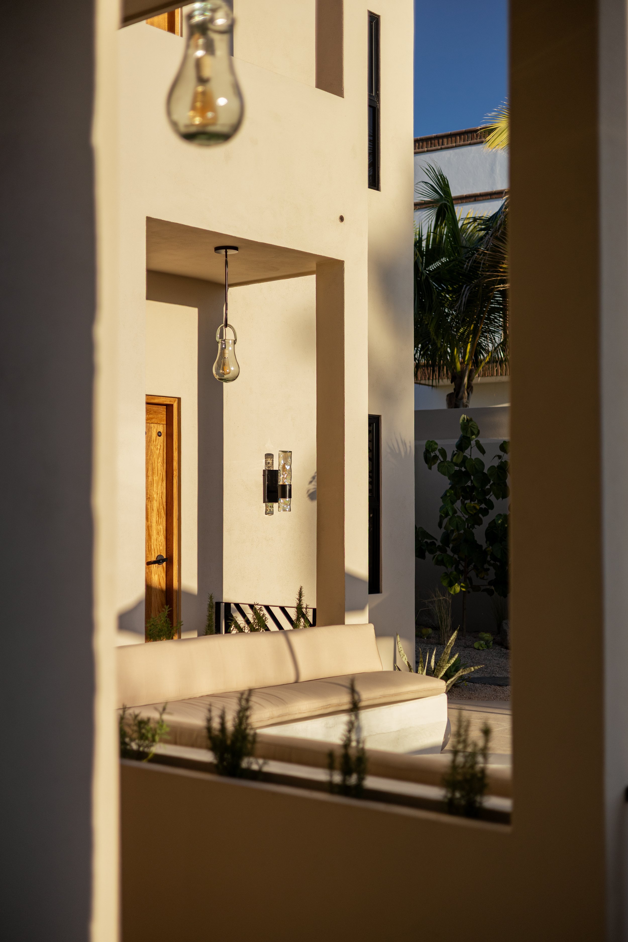 Outside view of a modern house with hanging light bulbs, a beige outdoor sofa, and tropical plants, taken at sunset.