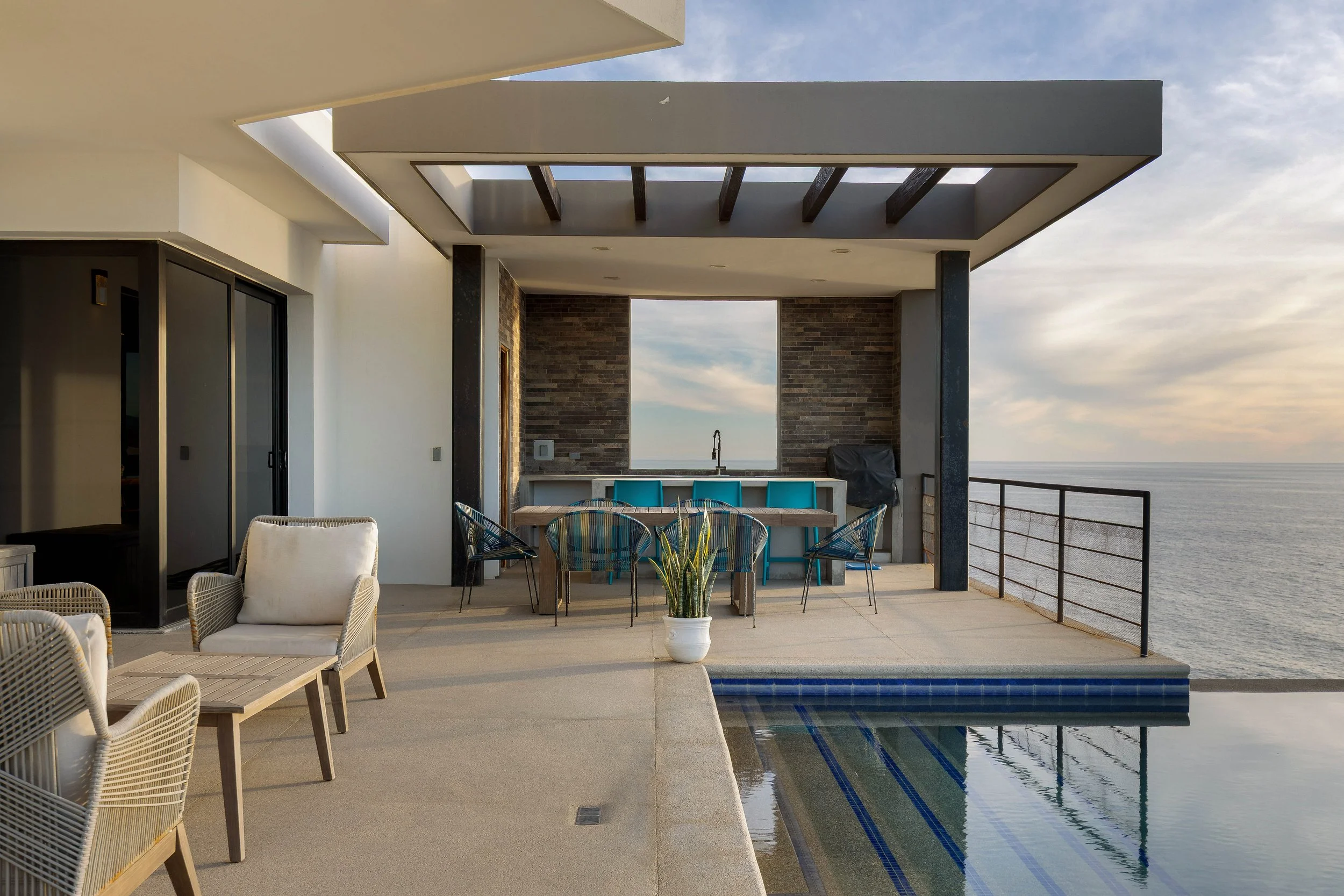 Modern outdoor patio with pool overlooking ocean, featuring seating, dining area, and bar under a partly open roof with sky view.