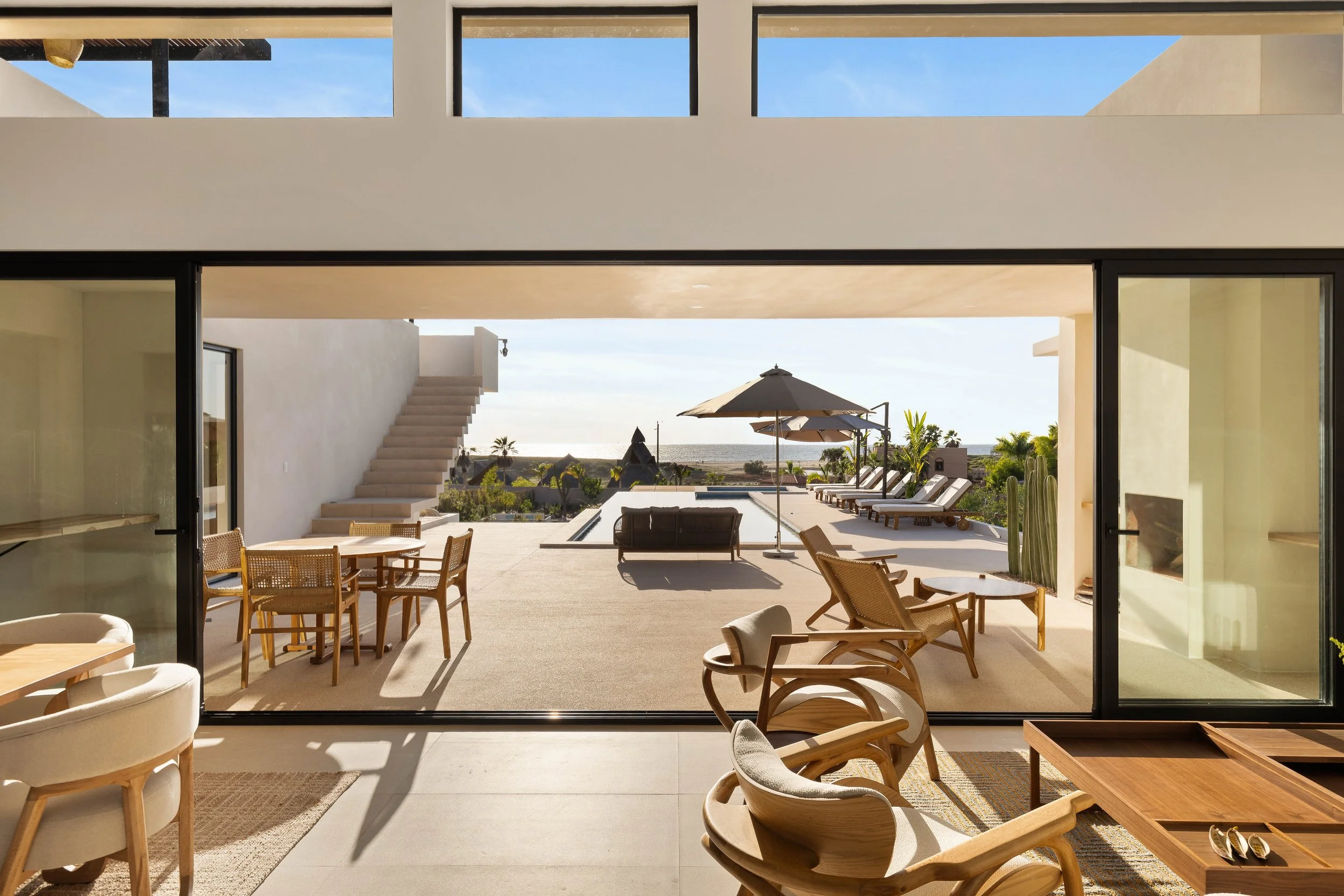 Indoor view of a modern house with open sliding glass doors leading to a patio with outdoor furniture, umbrellas, and a view of a desert landscape and ocean beyond.