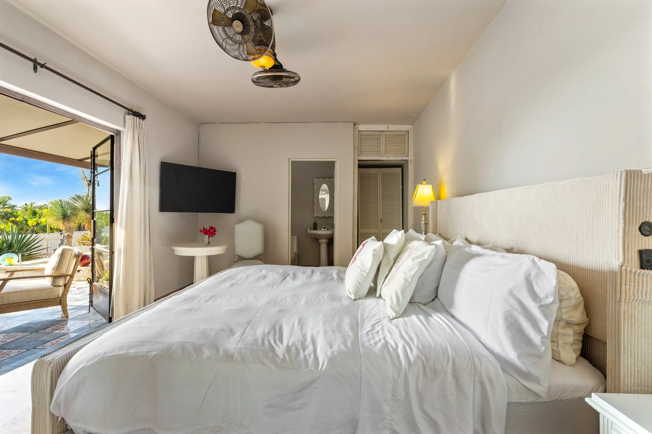 A bedroom with a large bed, white pillows, a lamp on the nightstand, a yellowish headboard, a small bathroom with a sink and mirror, an open balcony door with outdoor seating, and a wall-mounted TV.