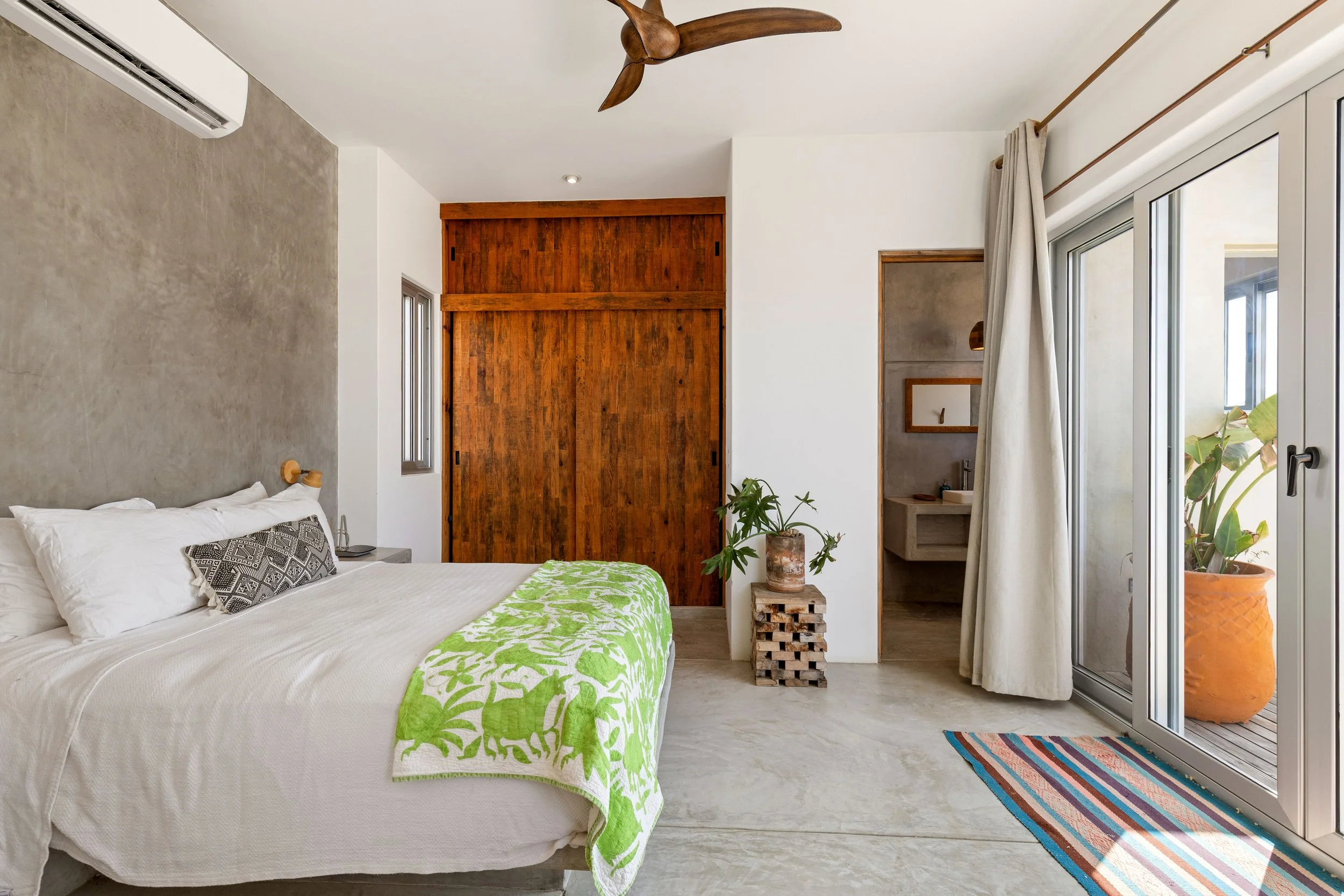 Modern bedroom with a bed covered in white linens and a green patterned blanket, wooden sliding closet doors, a small bathroom with a sink inside, and a large sliding glass door with cream curtains leading to a balcony with potted plants.