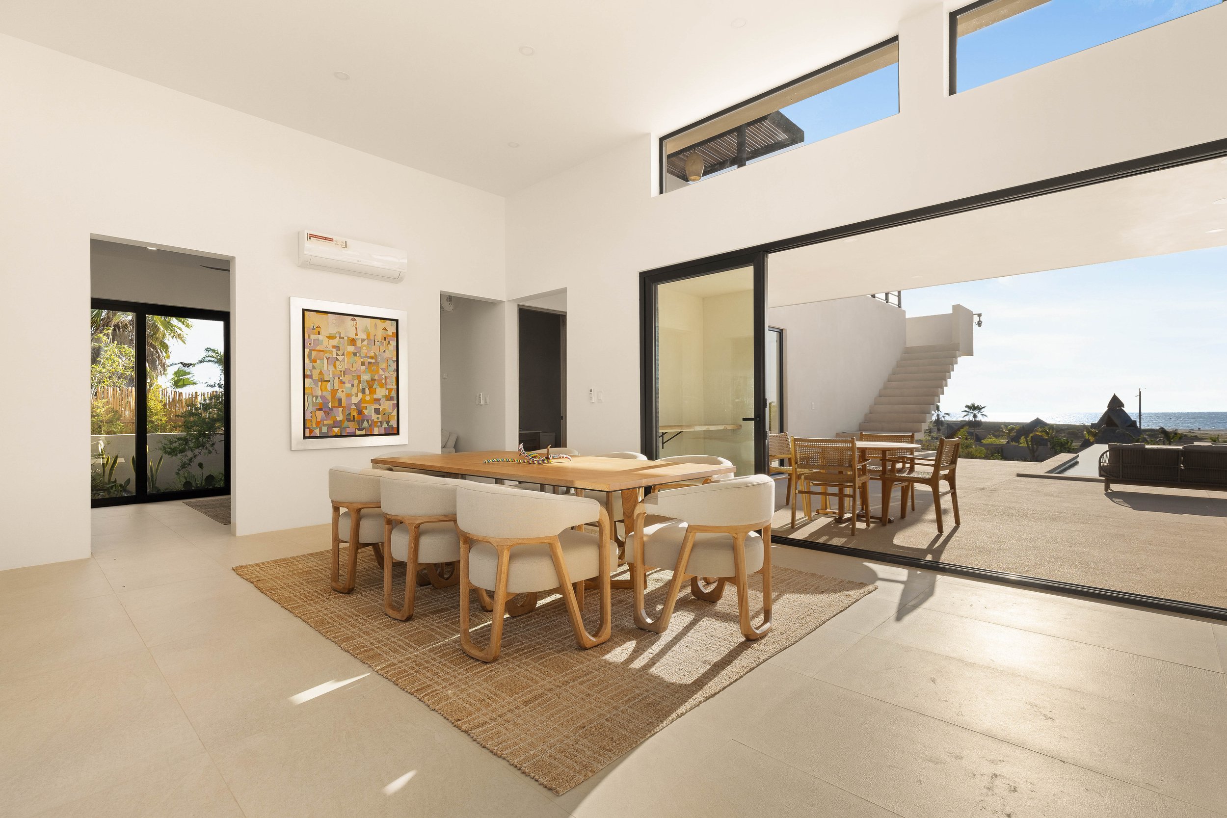 Modern dining area with a wooden table and beige chairs facing a large sliding glass door to an outdoor patio with ocean view.