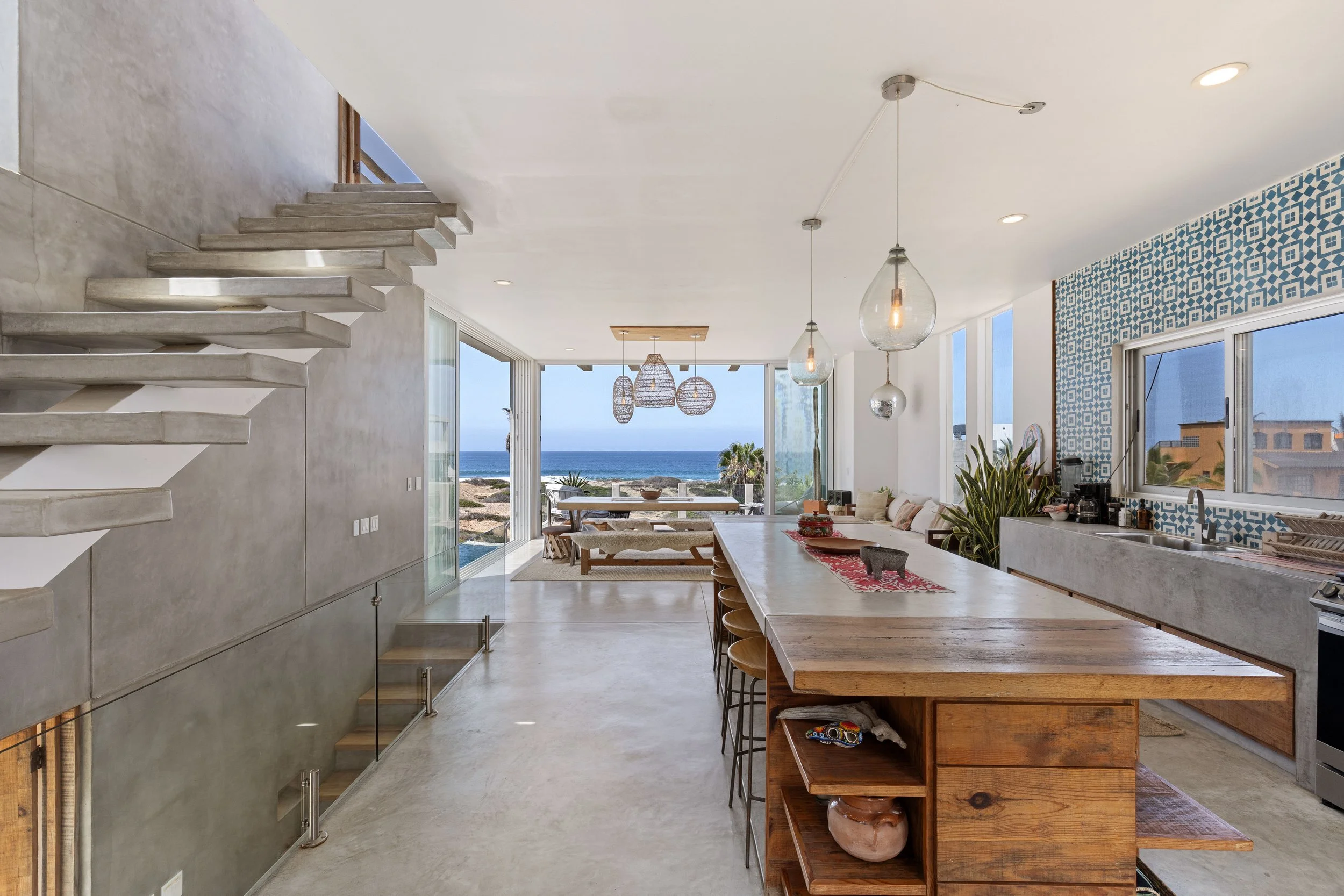 Modern open-concept kitchen and dining area with large windows and ocean view, featuring a long wooden kitchen island, pendant lights, patterned blue tile wall, and sliding glass doors leading outside.